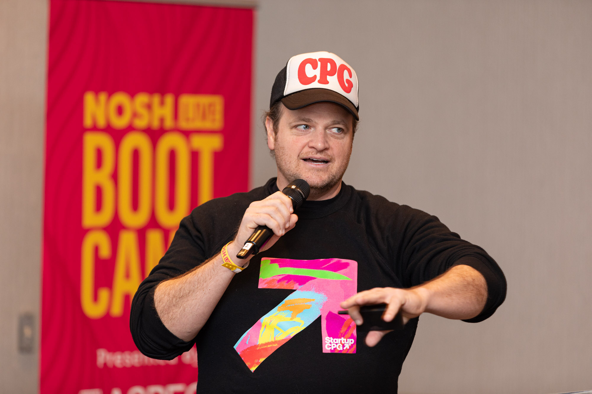 A man in a CPG hat and a black sweatshirt with a colorful logo speaking into a microphone in front of a Boot Camp banner.