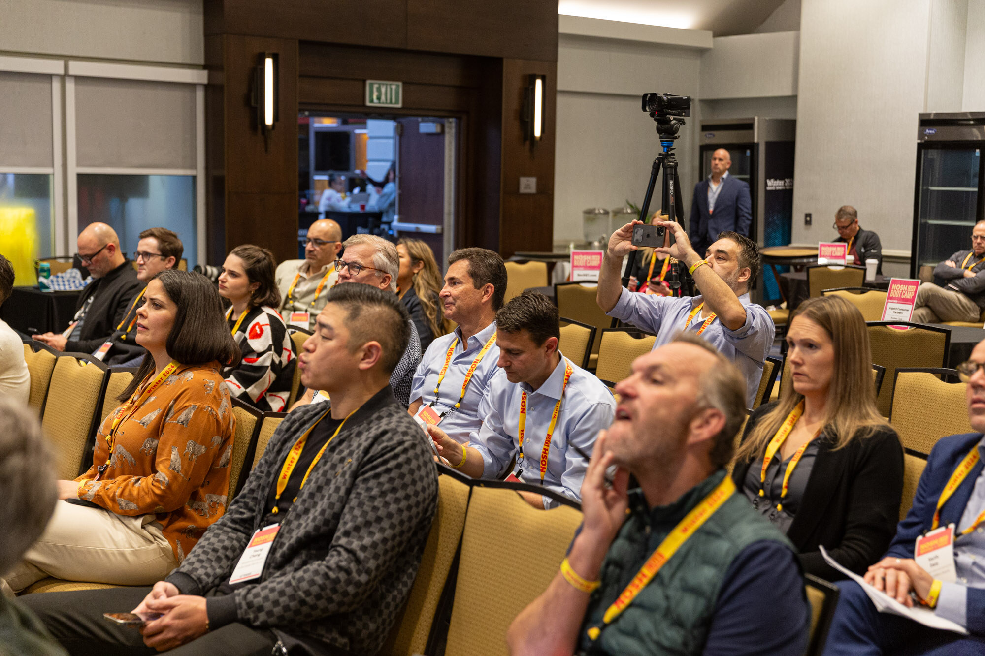 Rows of seated attendees in a hotel meeting room, some taking notes, one man looking up at the presenter.