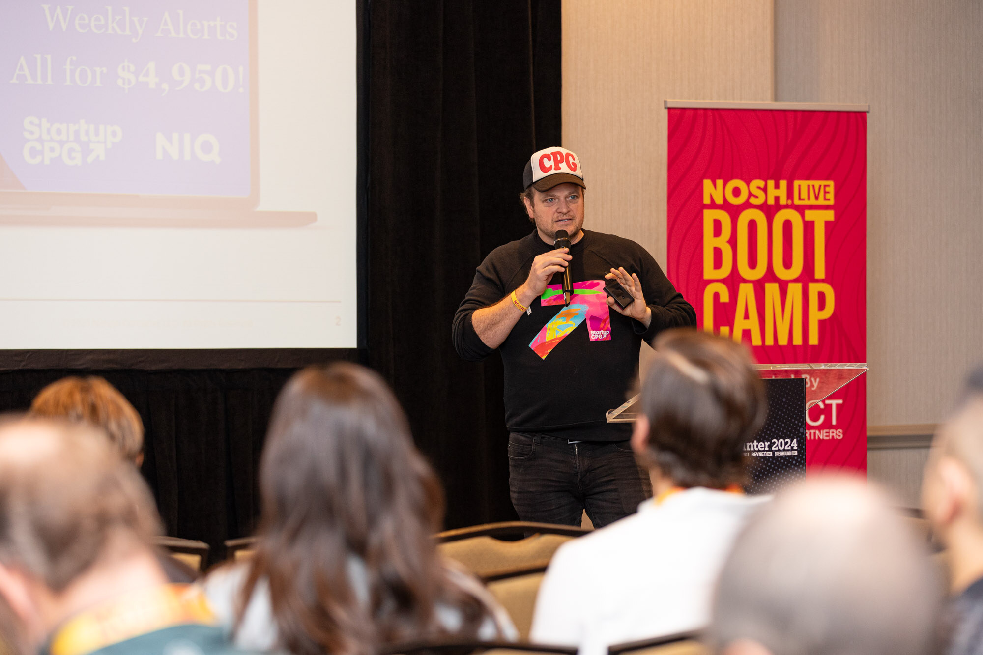 View over audience heads of a presenter beside a screen reading Weekly Alerts All for $4,950 with StartupCPG and NIQ logos.