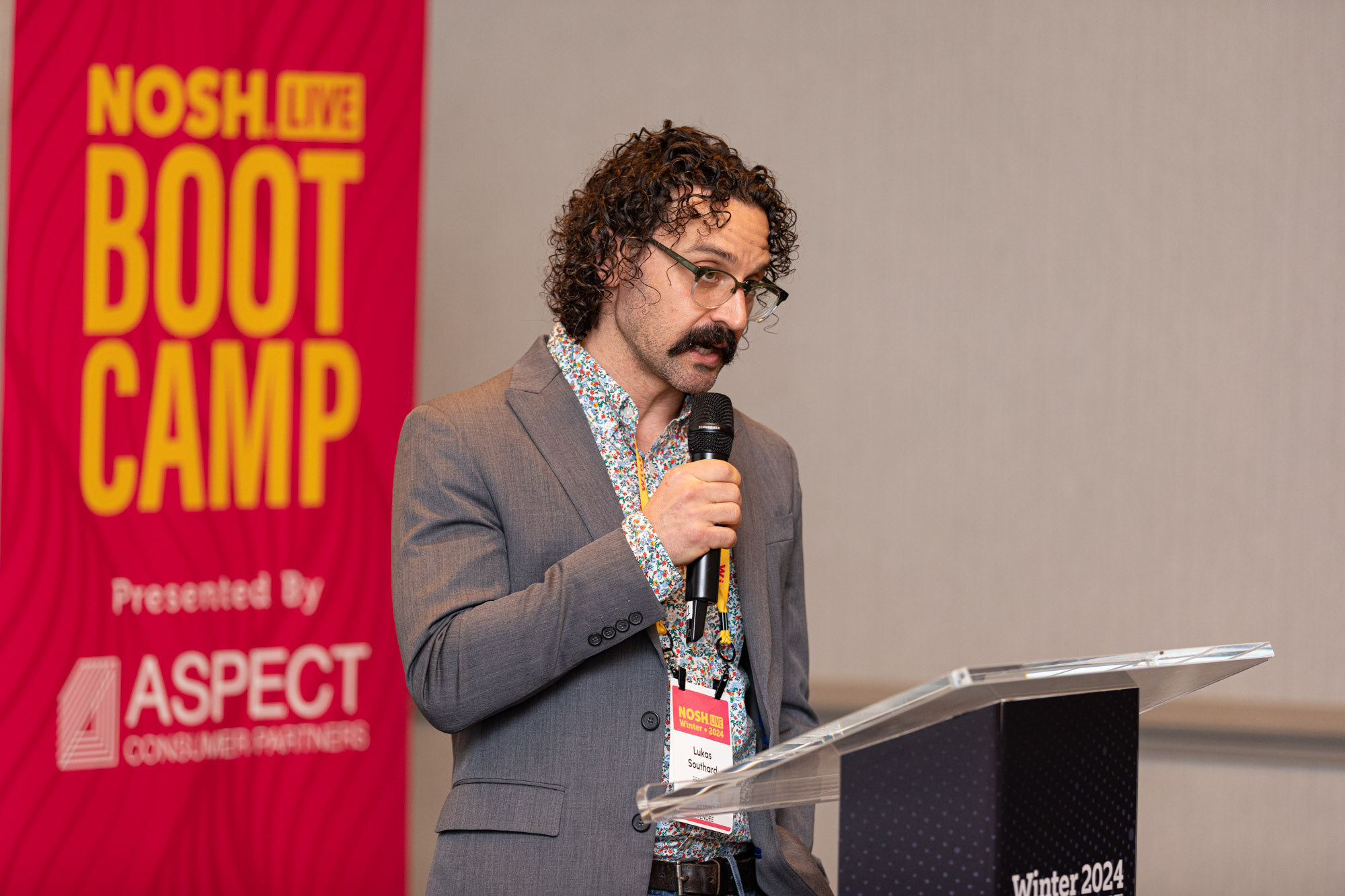 A man with curly hair and a mustache in a gray blazer speaking beside a red Nosh Live Boot Camp banner.