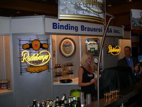 A blonde woman stands at a bar in a booth with Binding Brauerei signage and illuminated Radeberger signs, with bottles in the foreground and a man in a suit behind the bar.