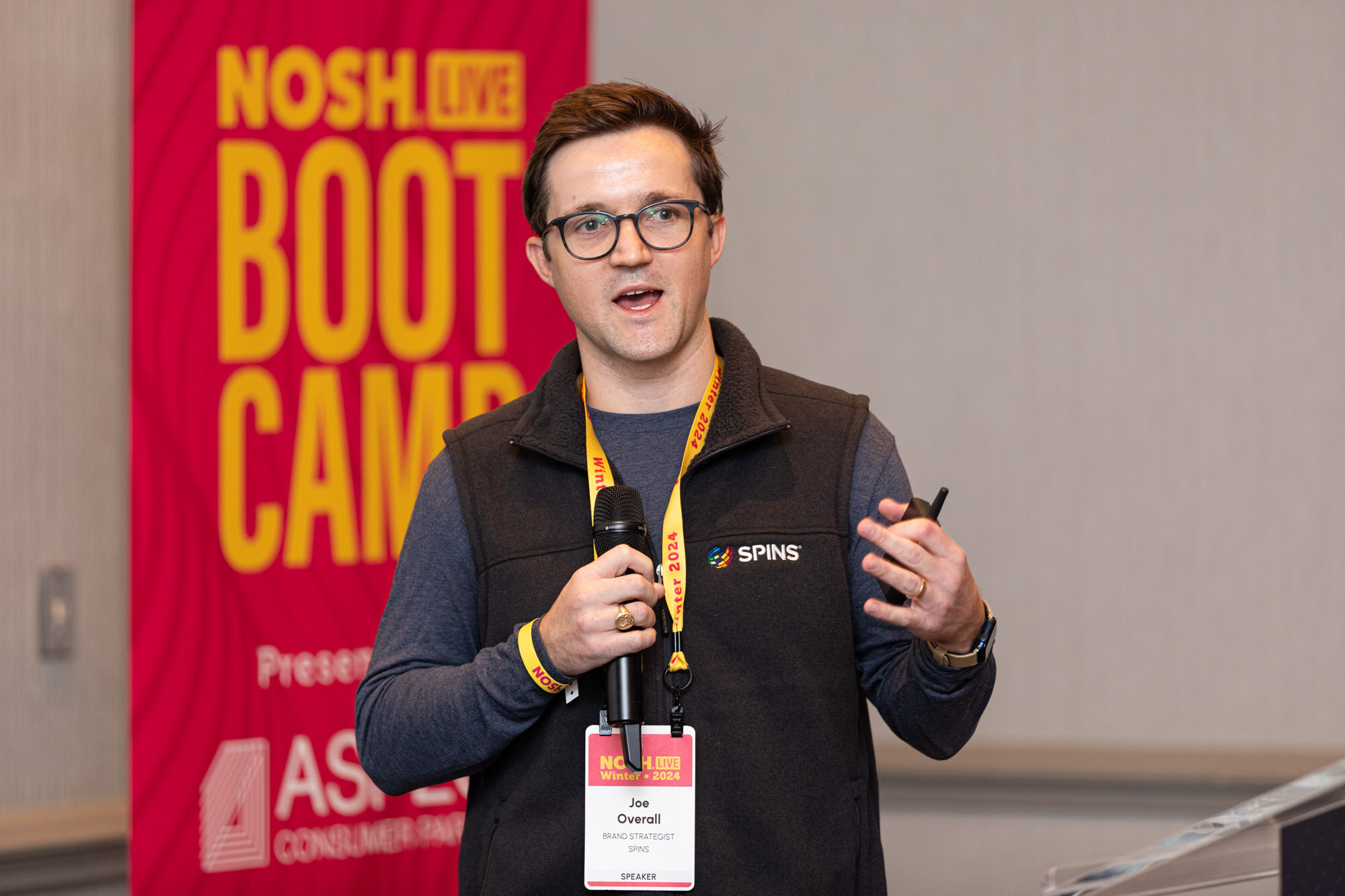 A man in dark glasses and a black SPINS vest holding a microphone and clicker in front of a Boot Camp banner.
