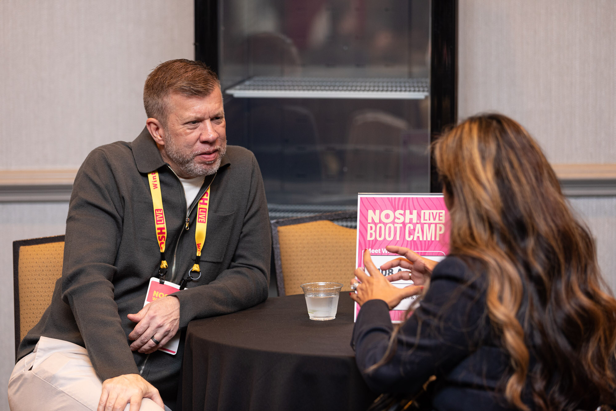 A man in a gray zip cardigan with a lanyard talking with a woman at a table with a pink Nosh Live Boot Camp sign.