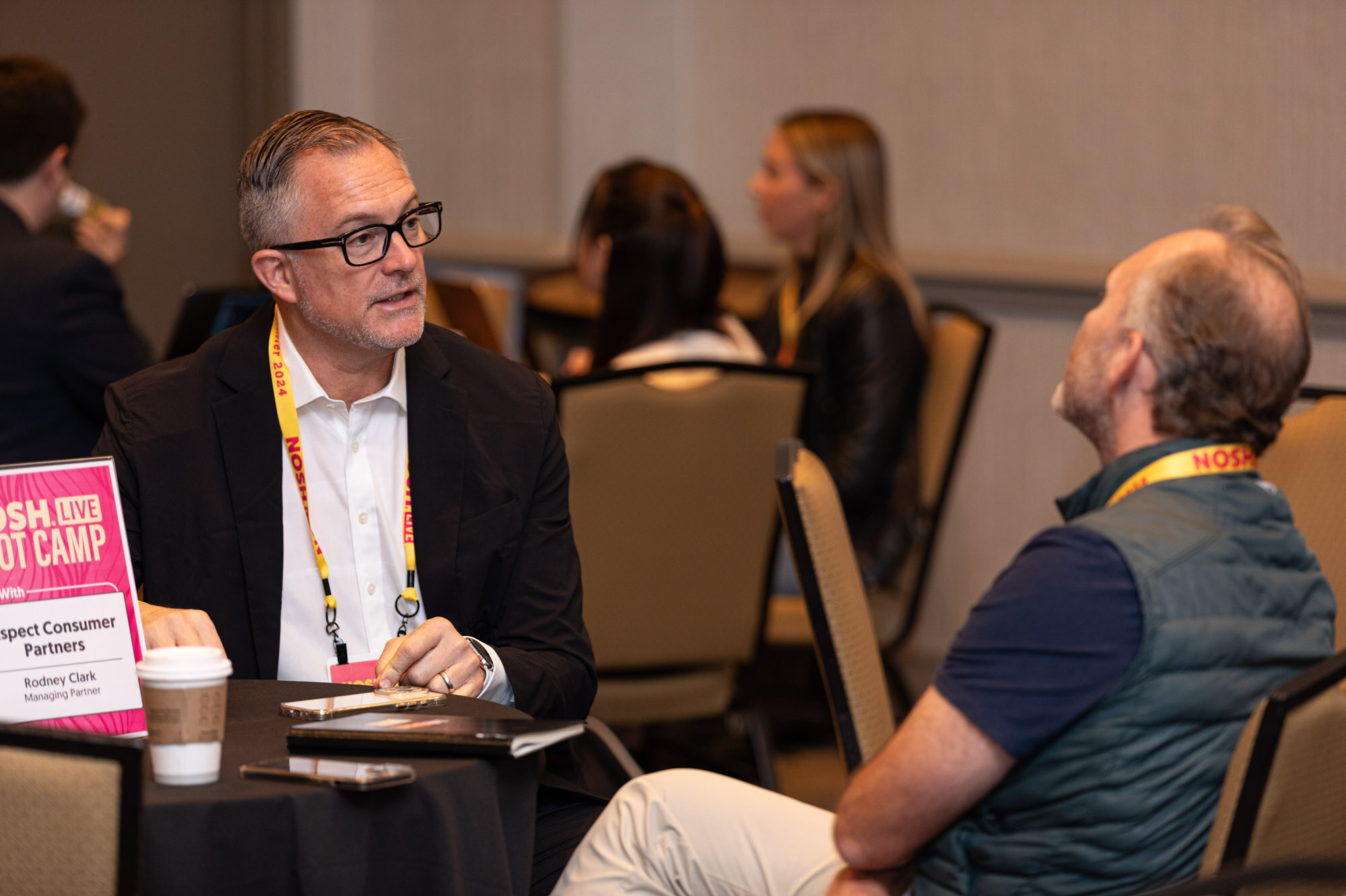 Two men talking at a round table with a pink sign reading Nosh Live Boot Camp Host With Aspect Consumer Partners.