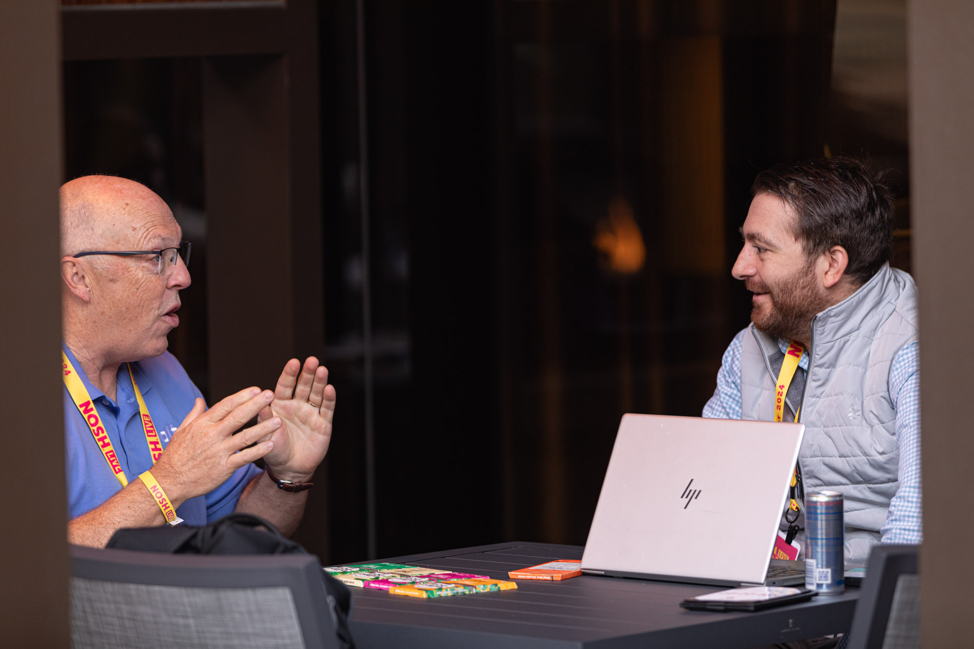 Two men talking across a table with a laptop in an outdoor lounge booth with a pink Nosh Live sign.