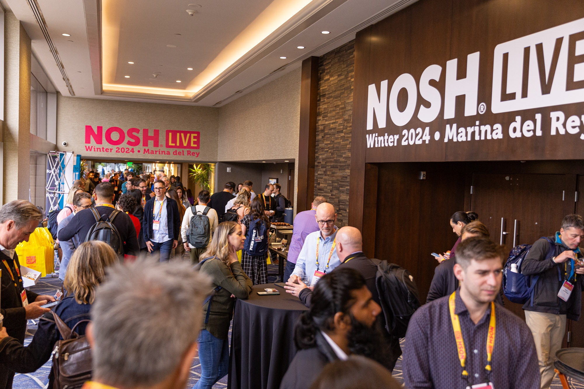Wide view of a crowded hallway with people chatting and a large Nosh Live sign on the far wall.