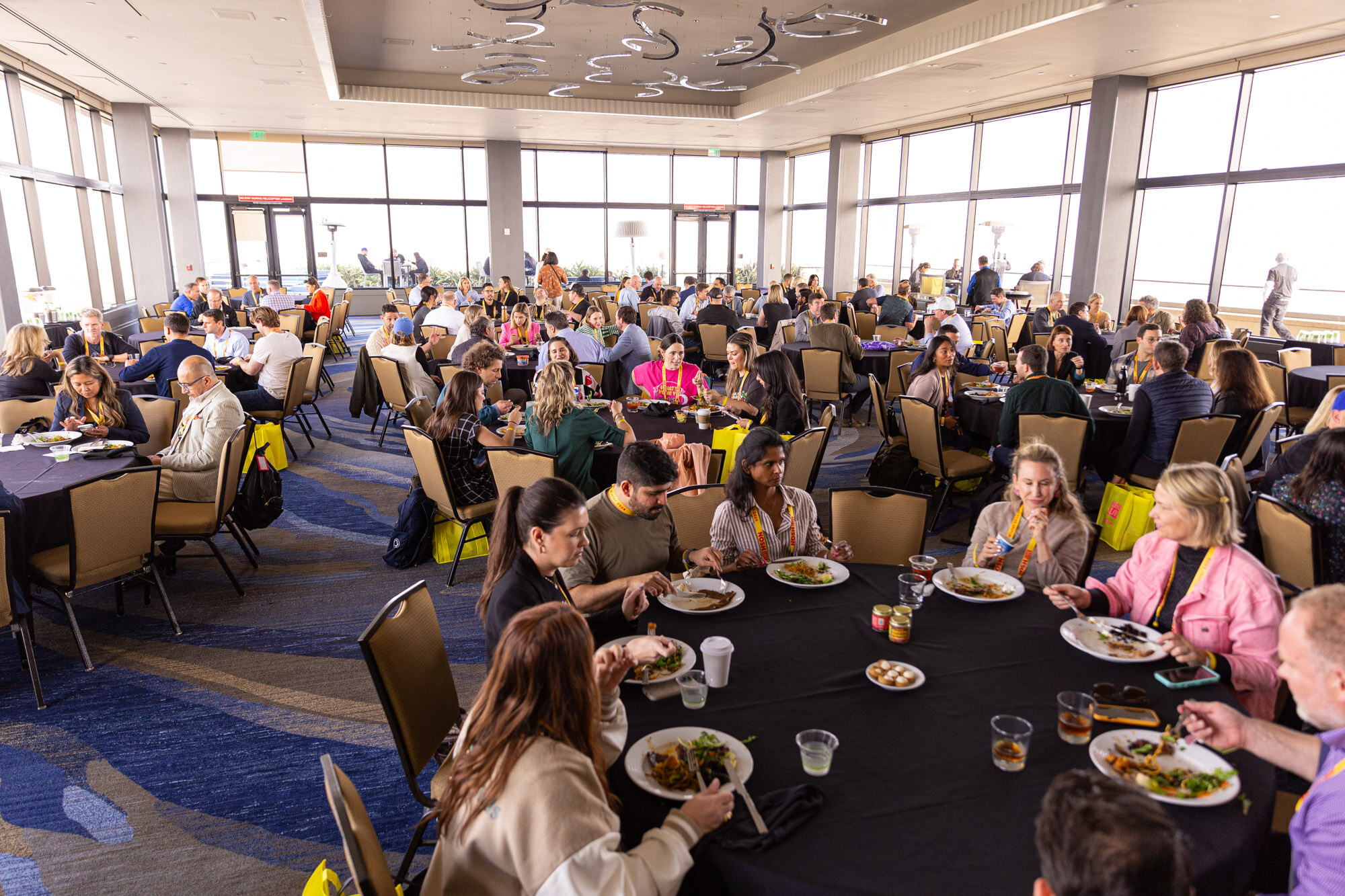 Wide view of a ballroom with many round tables where attendees sit eating.