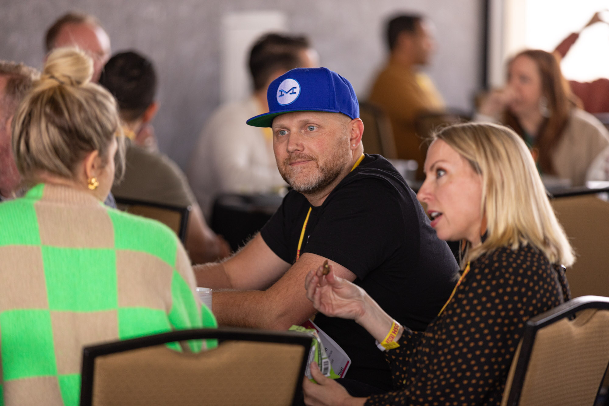 A man in a blue cap and black t-shirt and a blonde woman talking at a crowded ballroom table.