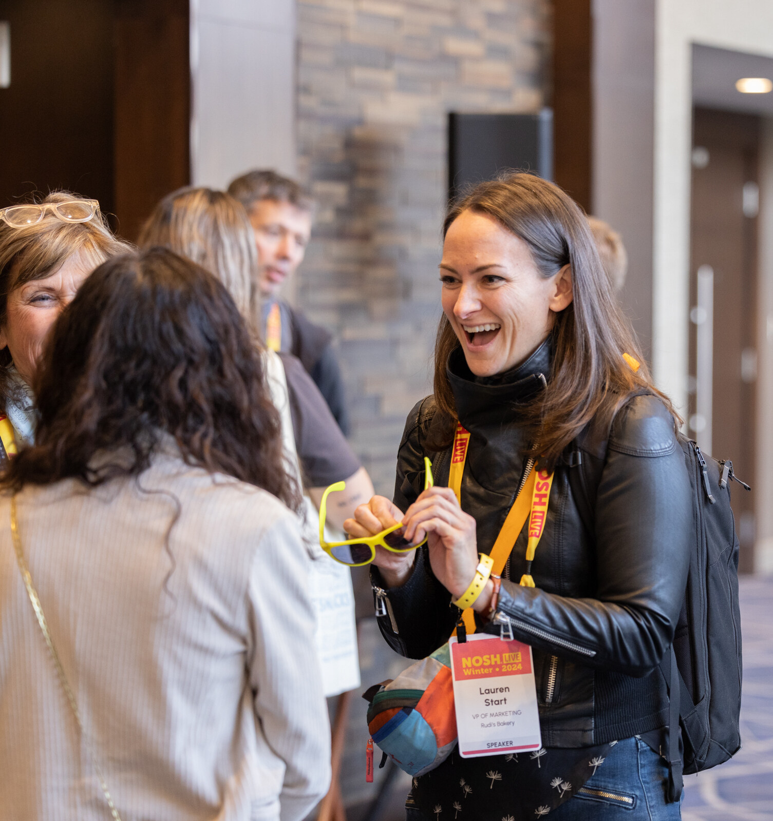 A woman in a black leather jacket with a lanyard laughing, holding yellow sunglasses in the hallway.