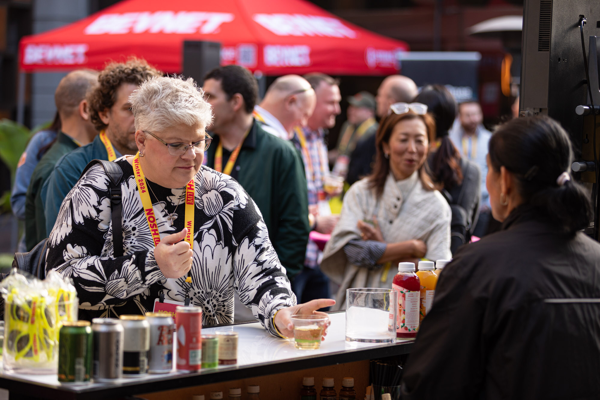 Attendees chatting at a bar with cans and bottles beside a red BevNET tent.