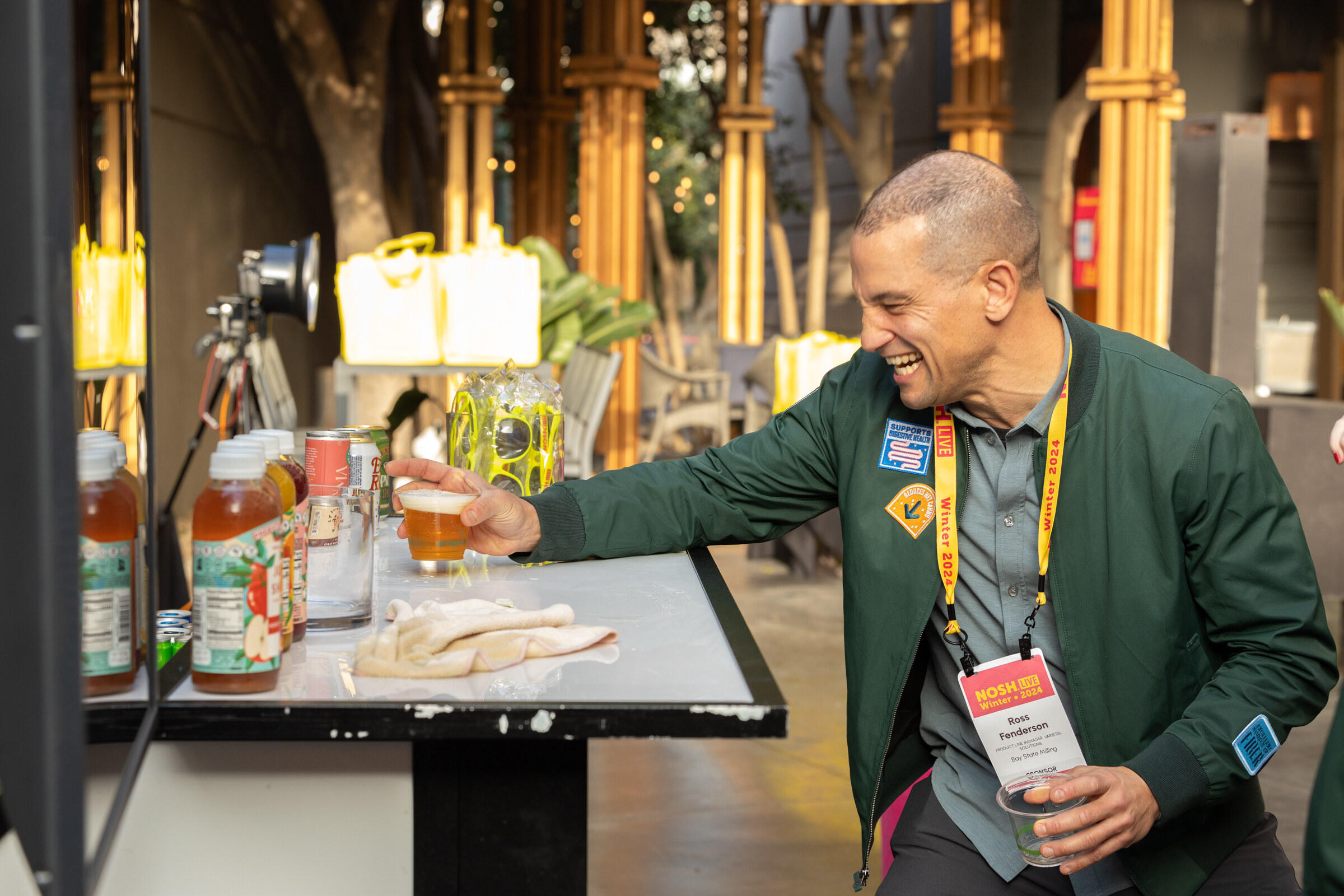 A man in a green jacket with a lanyard laughing and picking up a cup next to bottles on a table.