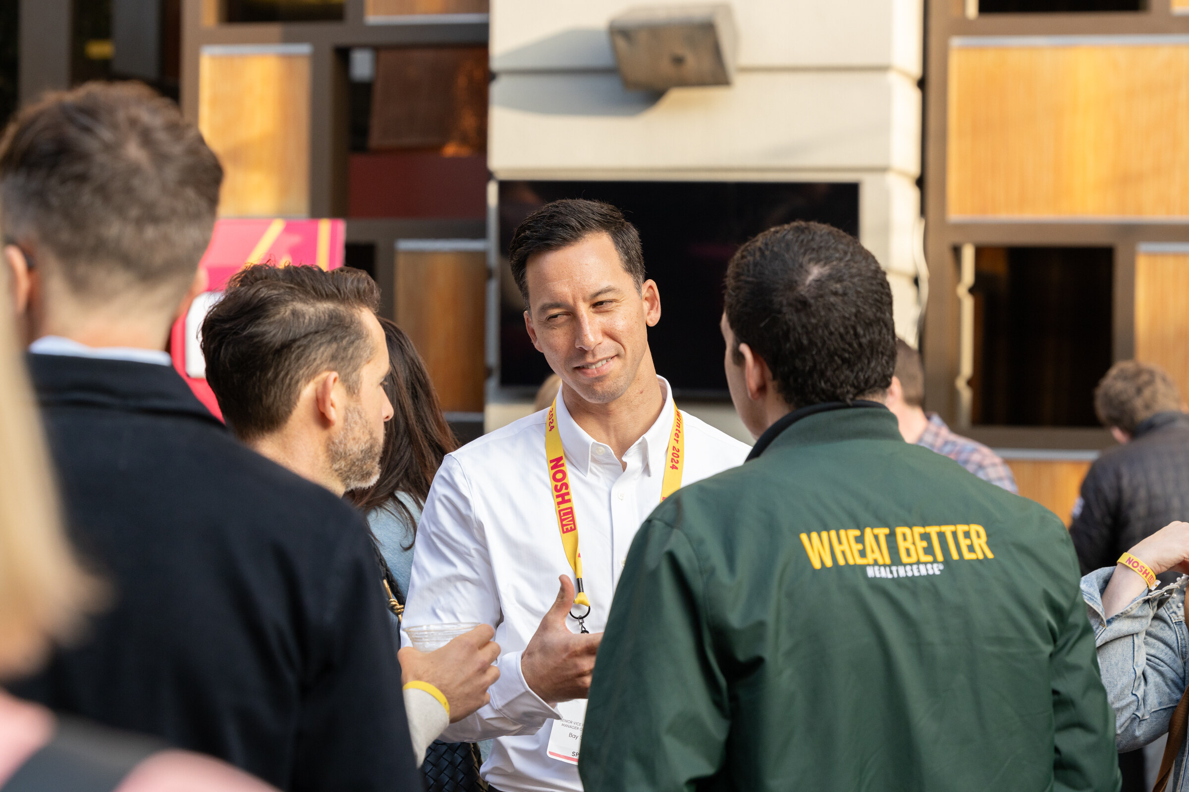 Several attendees in conversation outdoors, one wearing a green jacket with Wheat Better on the back.