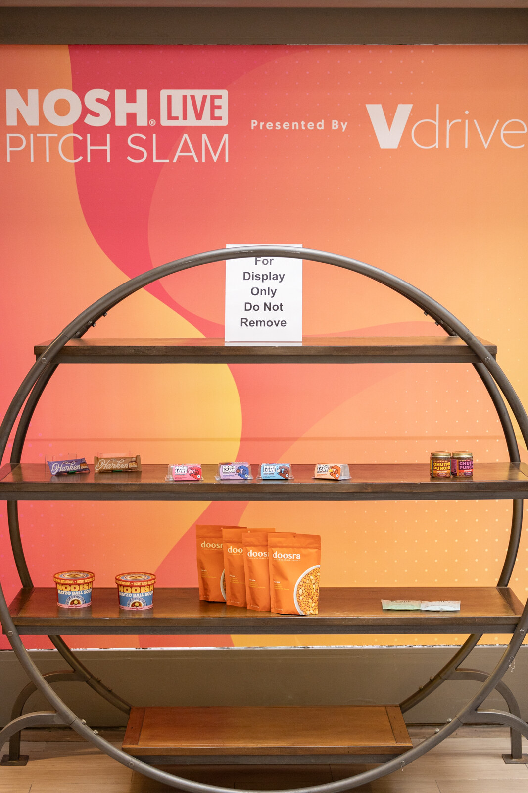 A circular shelf displaying product bags and jars below a sign reading Nosh Live Pitch Slam Presented by Vdriven.