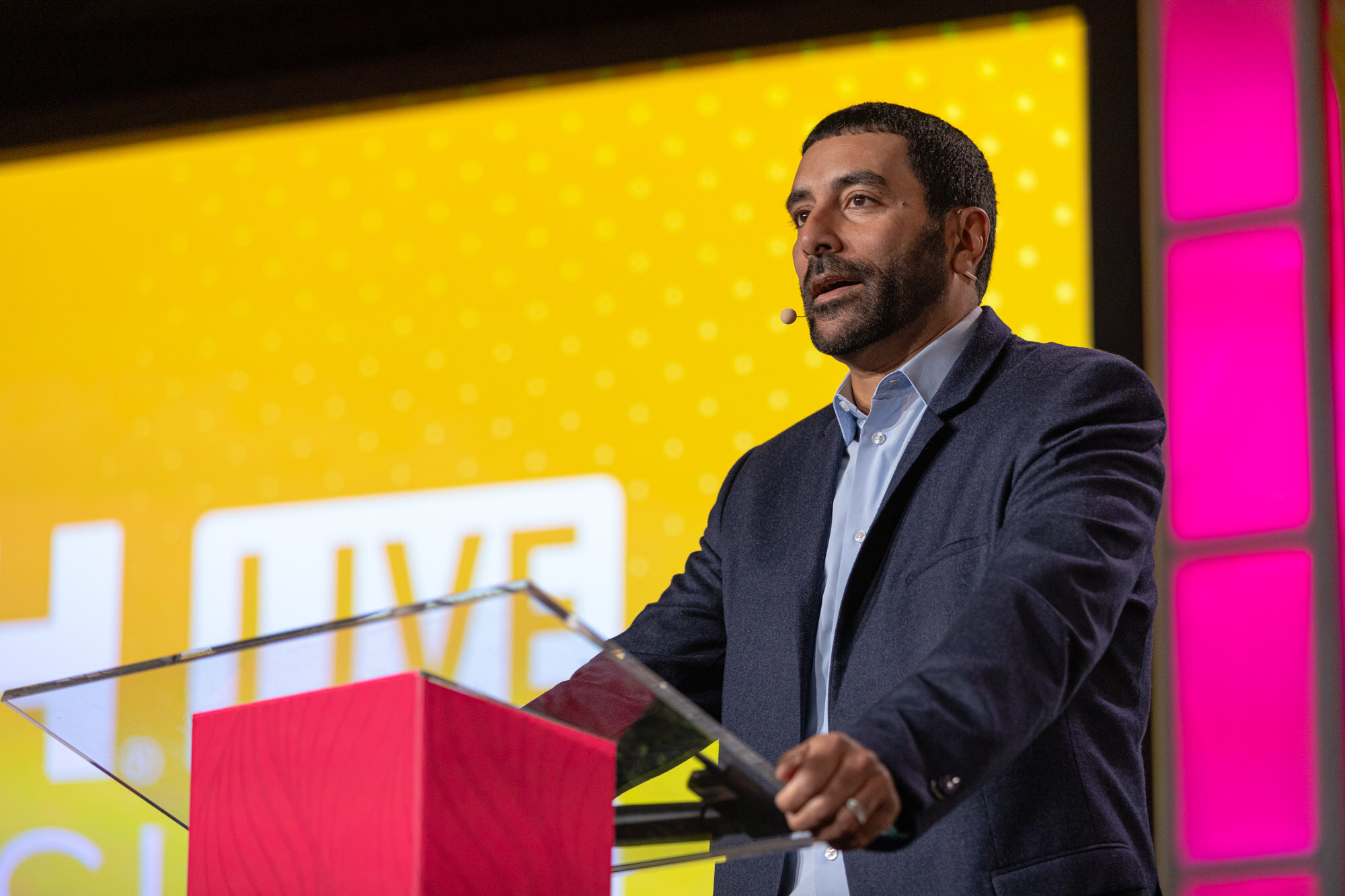 A bearded man in a navy blazer at a red Nosh Live podium speaking.