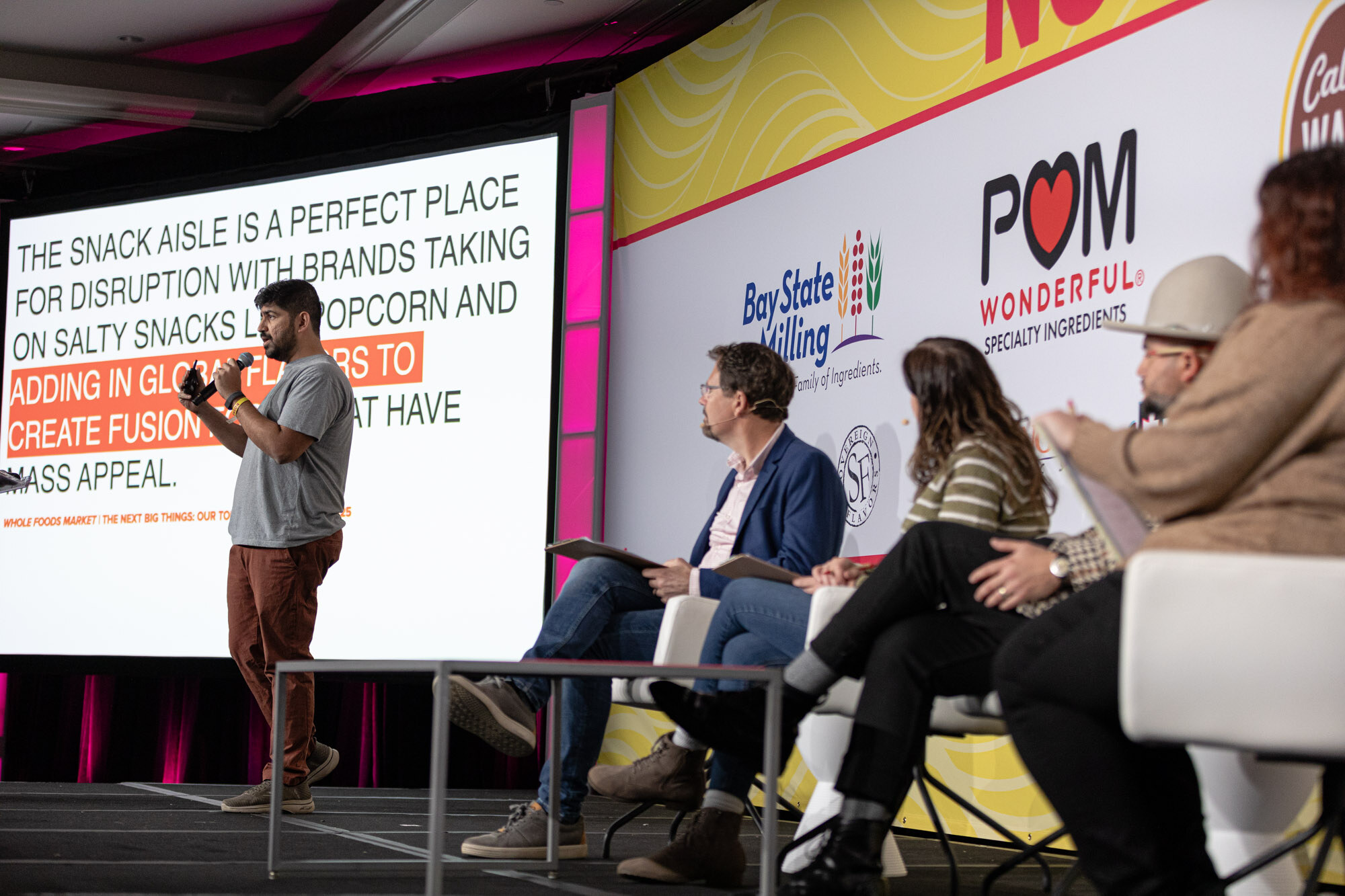 Wide view of a presenter at left, four seated judges at right, and a slide about the snack aisle on the screen.