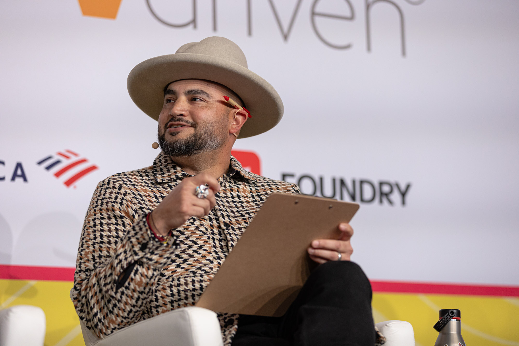 A man in a cream wide-brim hat and houndstooth shirt holding a clipboard, seated on stage.