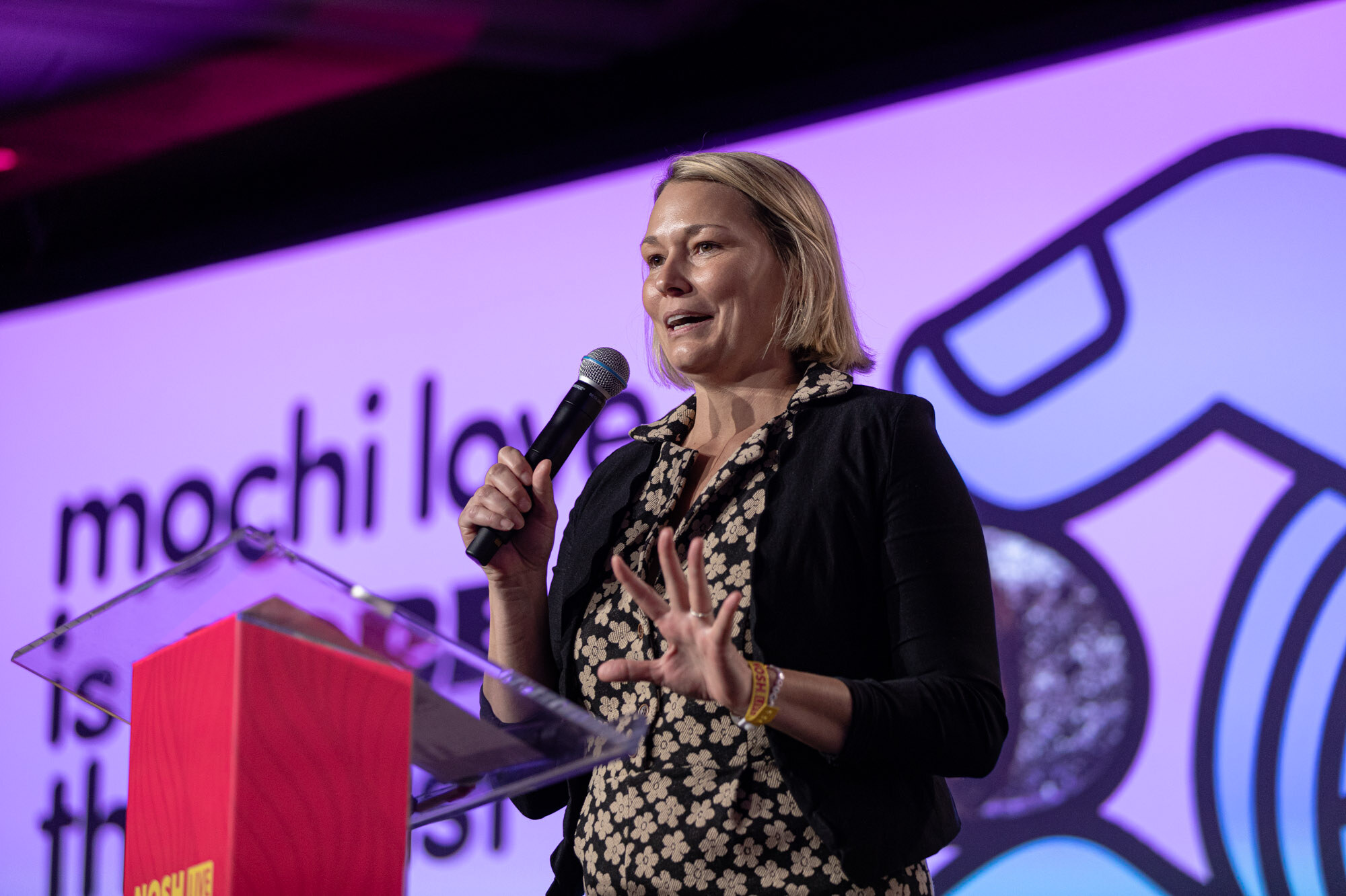 A woman with a blond bob and black blazer speaking on stage in front of a purple slide.