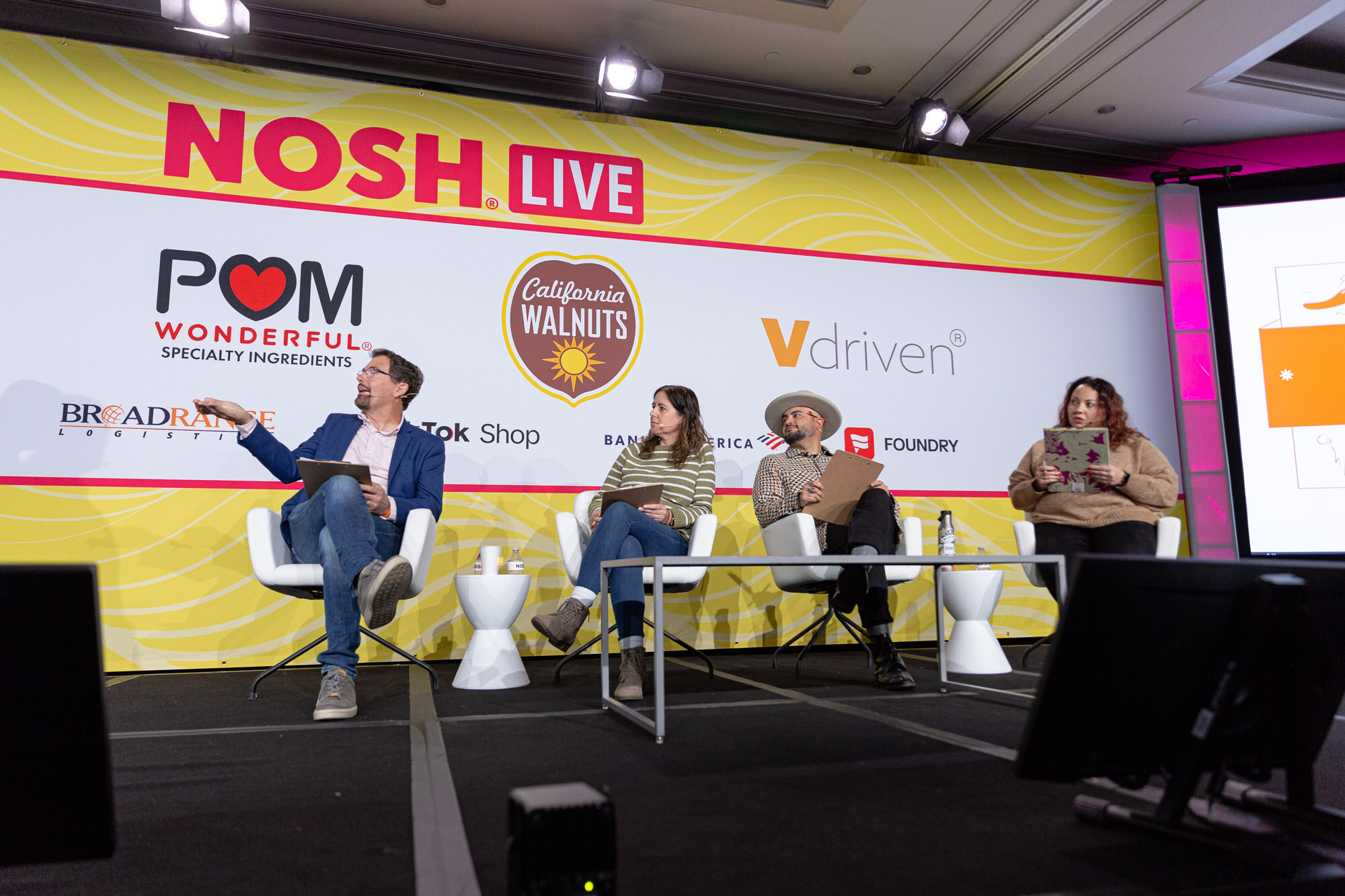 Wide view of four people seated in a row on a stage with a yellow Nosh Live backdrop.