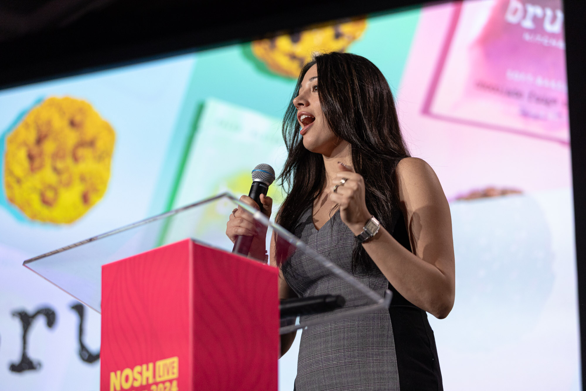 A woman with long dark hair in a gray dress speaking with a slide behind her.
