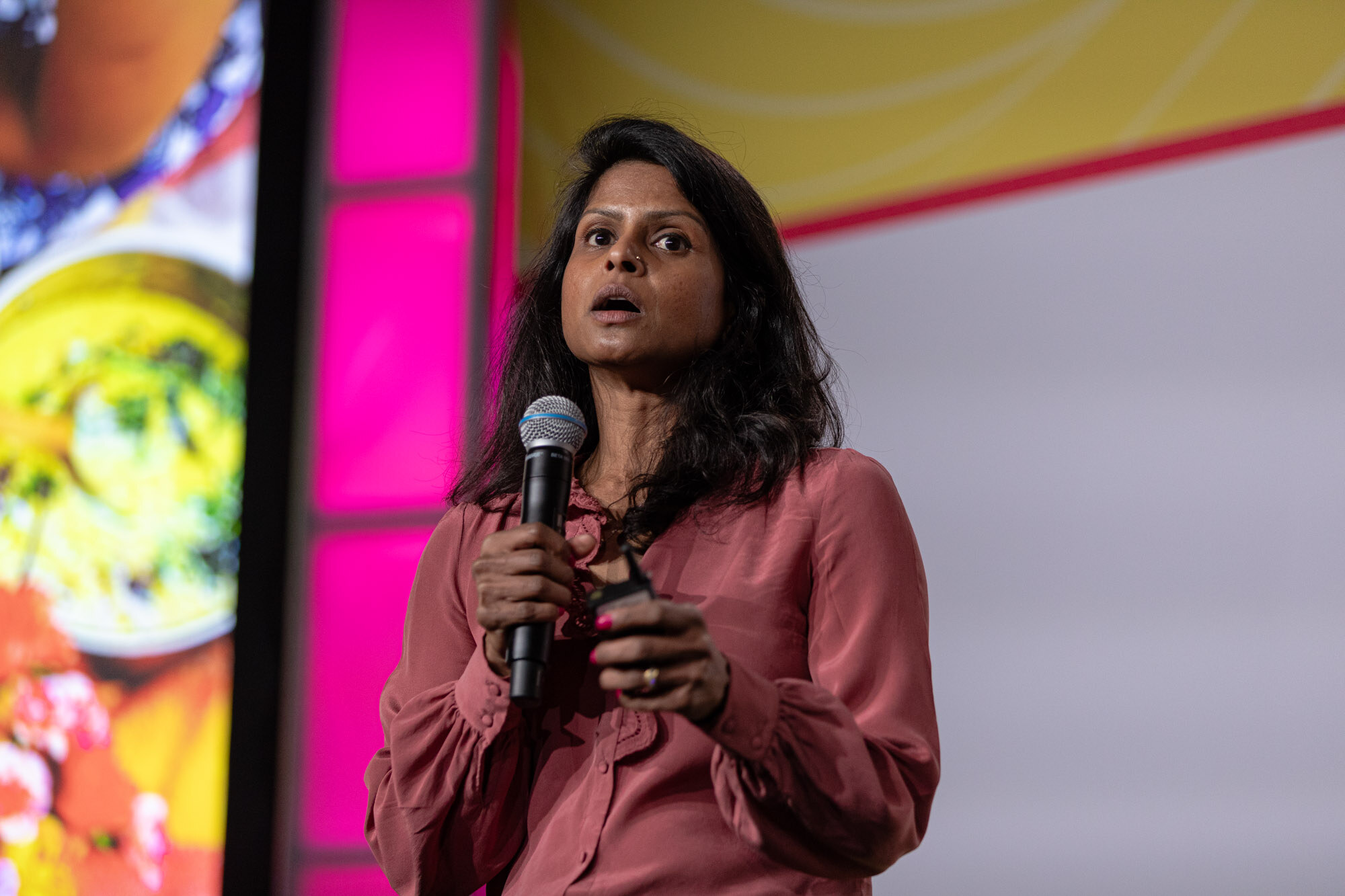 A woman in a rose-colored blouse and green pants speaking into a microphone in front of a Chutney Powder slide.
