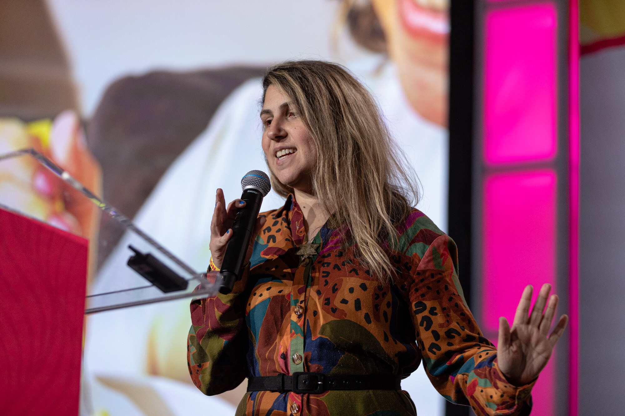 A woman with long wavy hair in a bright leopard-print shirt holding a microphone, with a slide behind her.