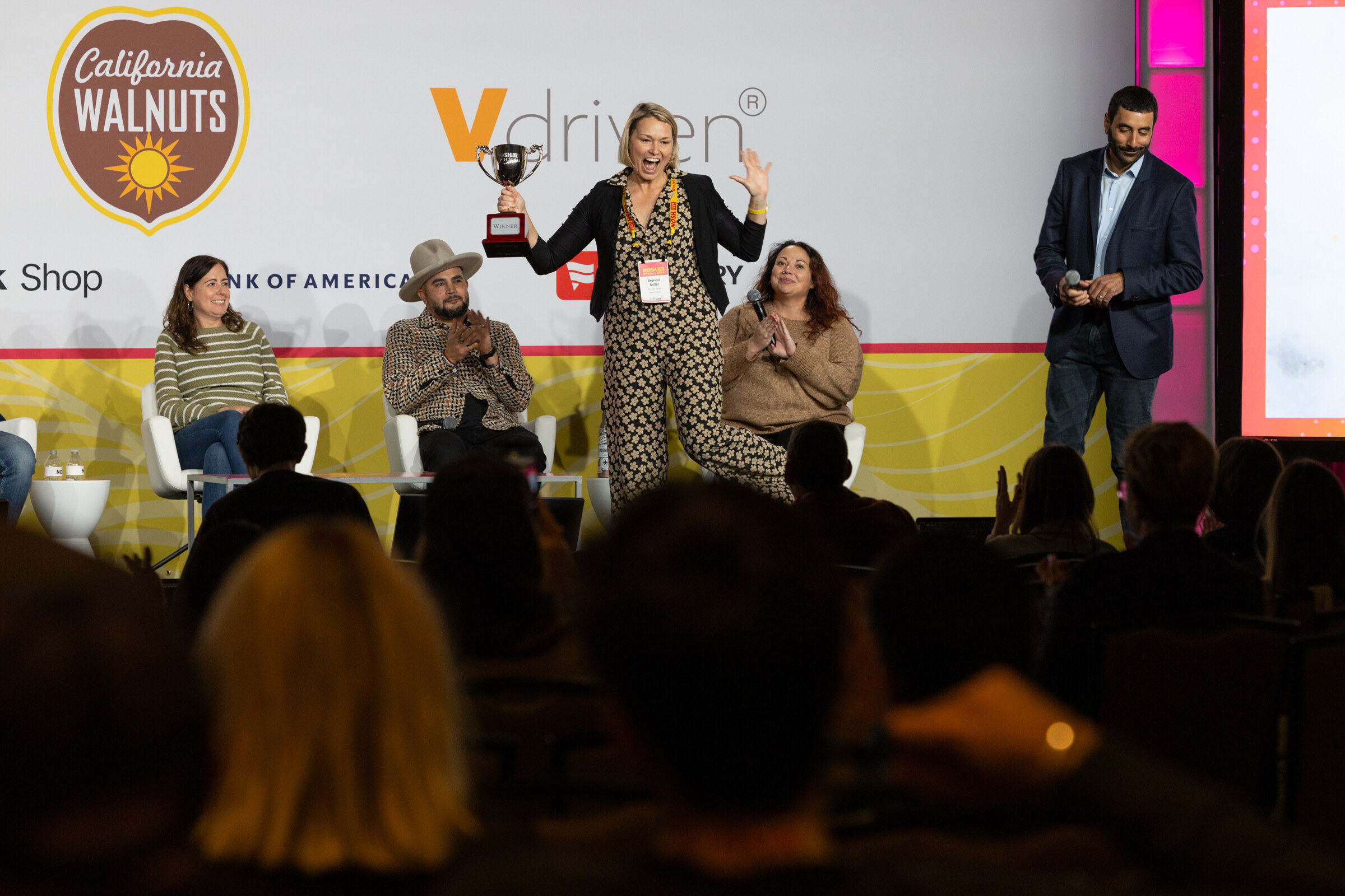 A woman in a black blazer and patterned jumpsuit holding a trophy overhead on stage next to a man in a dark suit and seated judges.