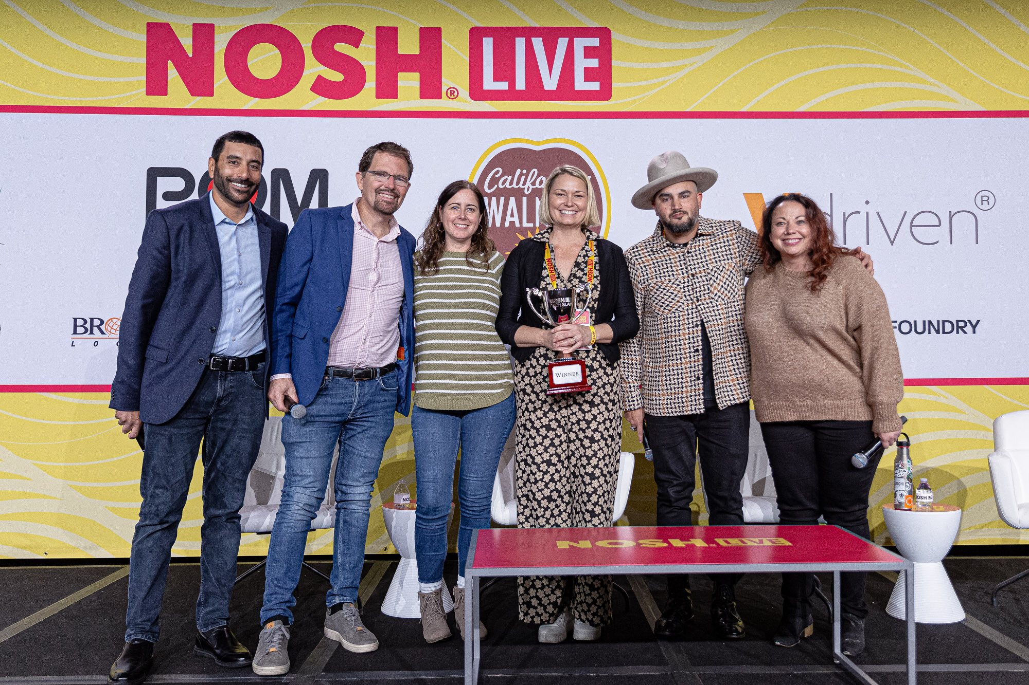 Six people standing together in front of the Nosh Live backdrop, one woman holding a trophy.