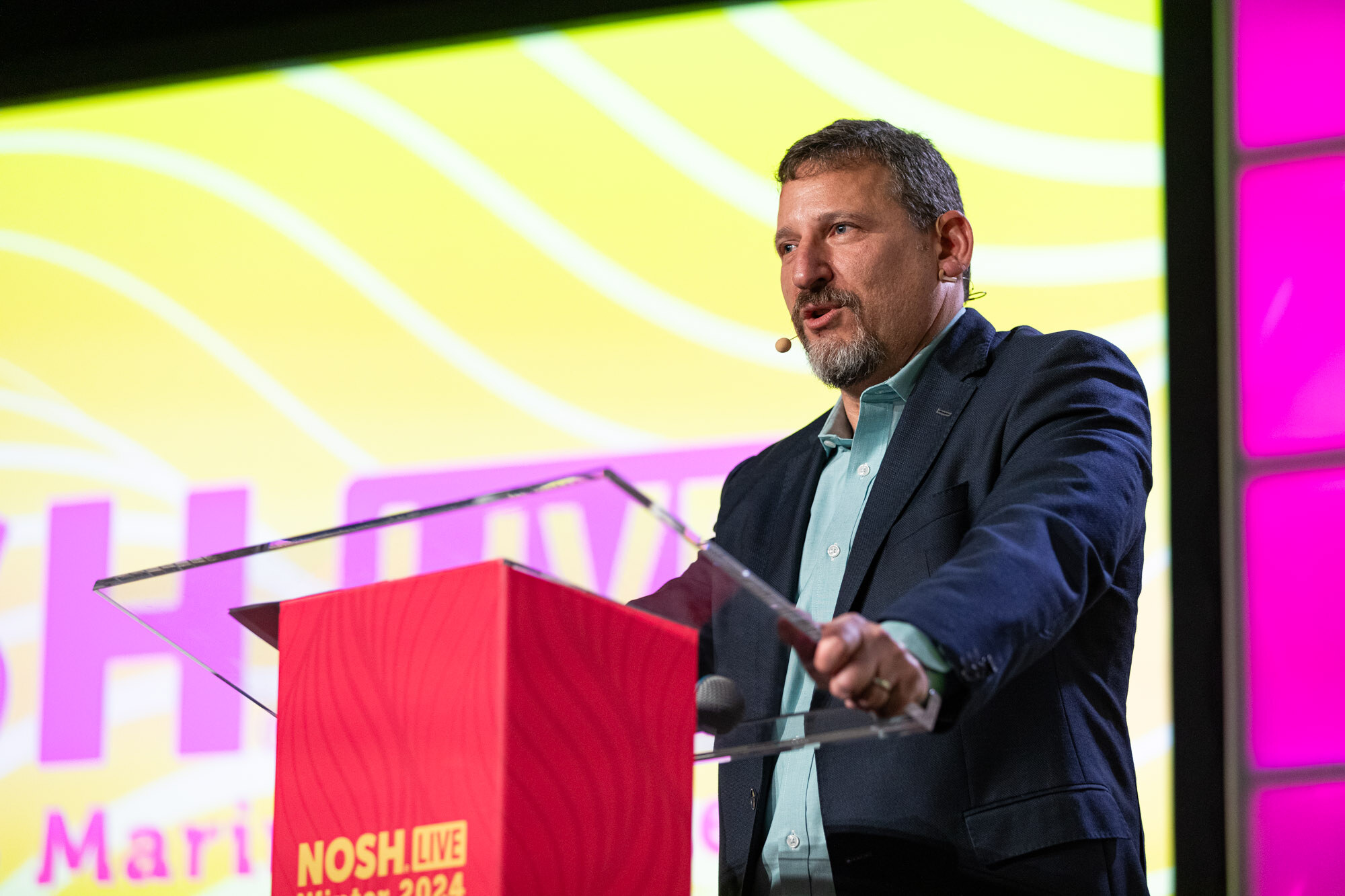 A man with a graying beard in a navy blazer and teal shirt speaking at a red podium in front of a yellow Nosh Live screen.