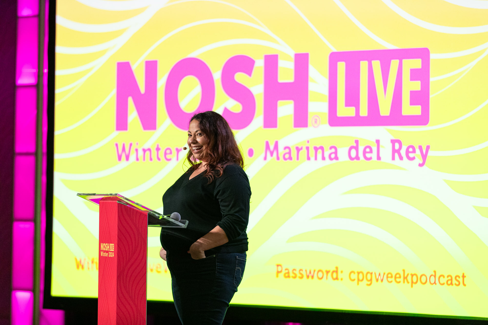 A woman in a black top standing at a podium beside a large yellow screen reading Nosh Live Winter 2024 Marina del Rey.
