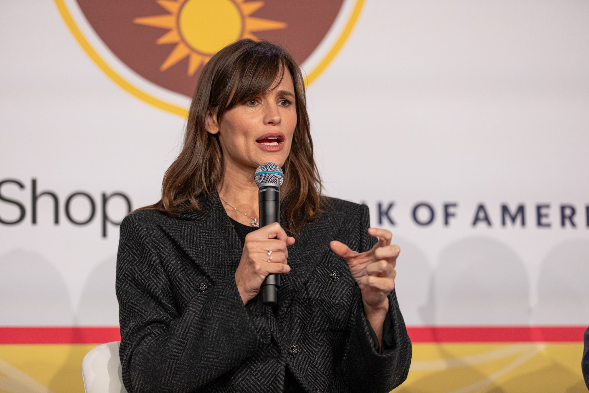 A woman with shoulder-length brown hair in a charcoal herringbone blazer speaking into a microphone in front of a sponsor wall.