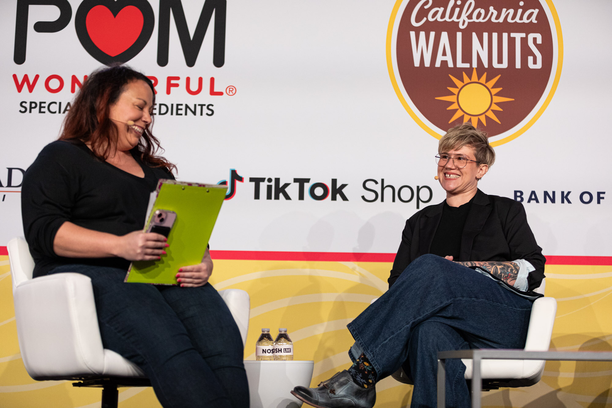 A woman holding a green folder sits beside a short-haired guest in glasses and a black blazer on a stage with sponsor logos behind them.