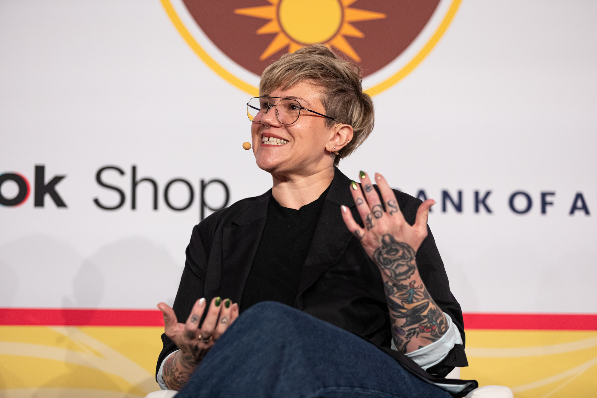 A woman with short blond hair, glasses and tattooed hands wearing a black blazer, gesturing while seated on stage.