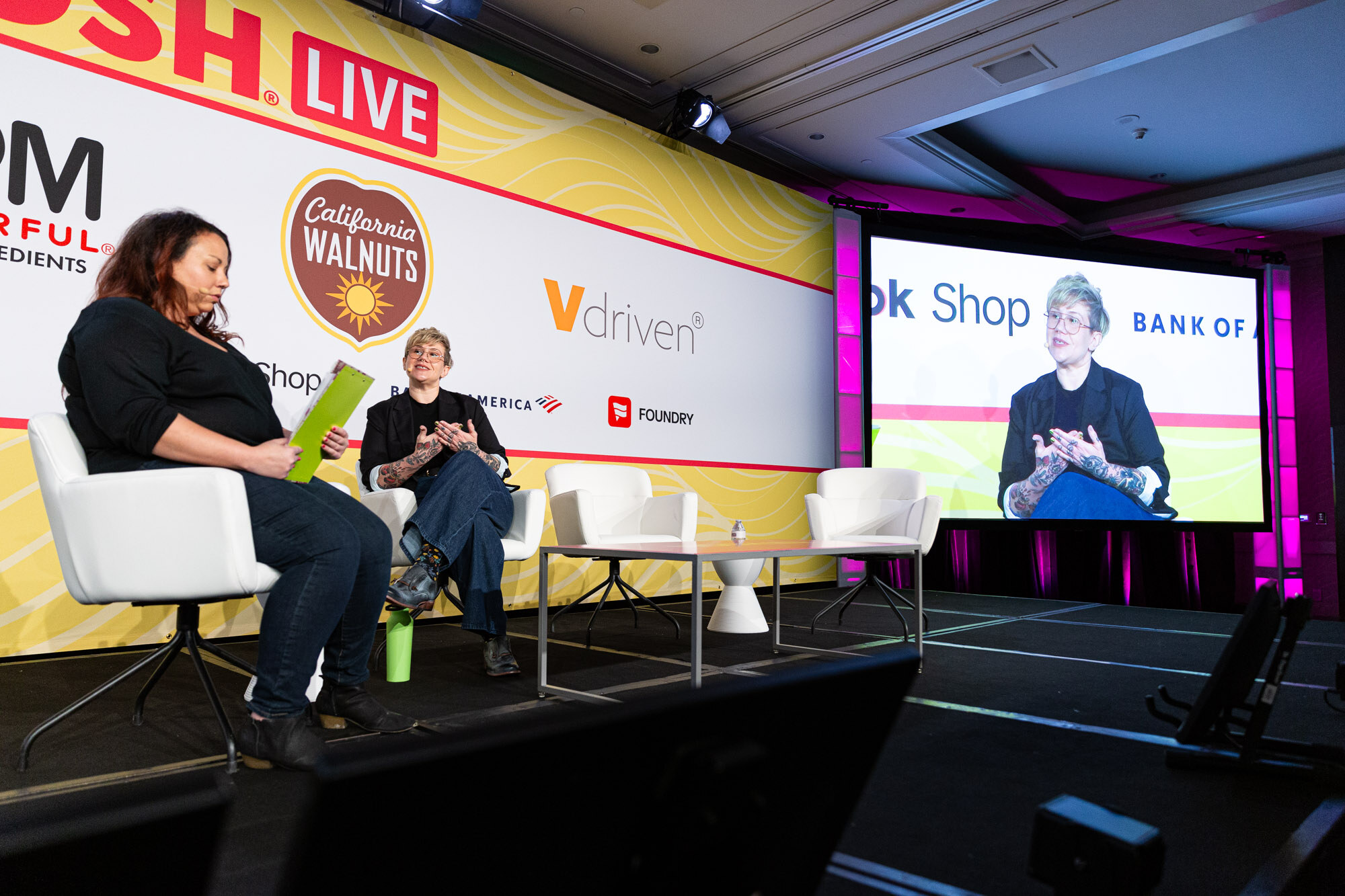 Wide view of two people seated on a stage with a large screen at the right showing a close-up of the guest.