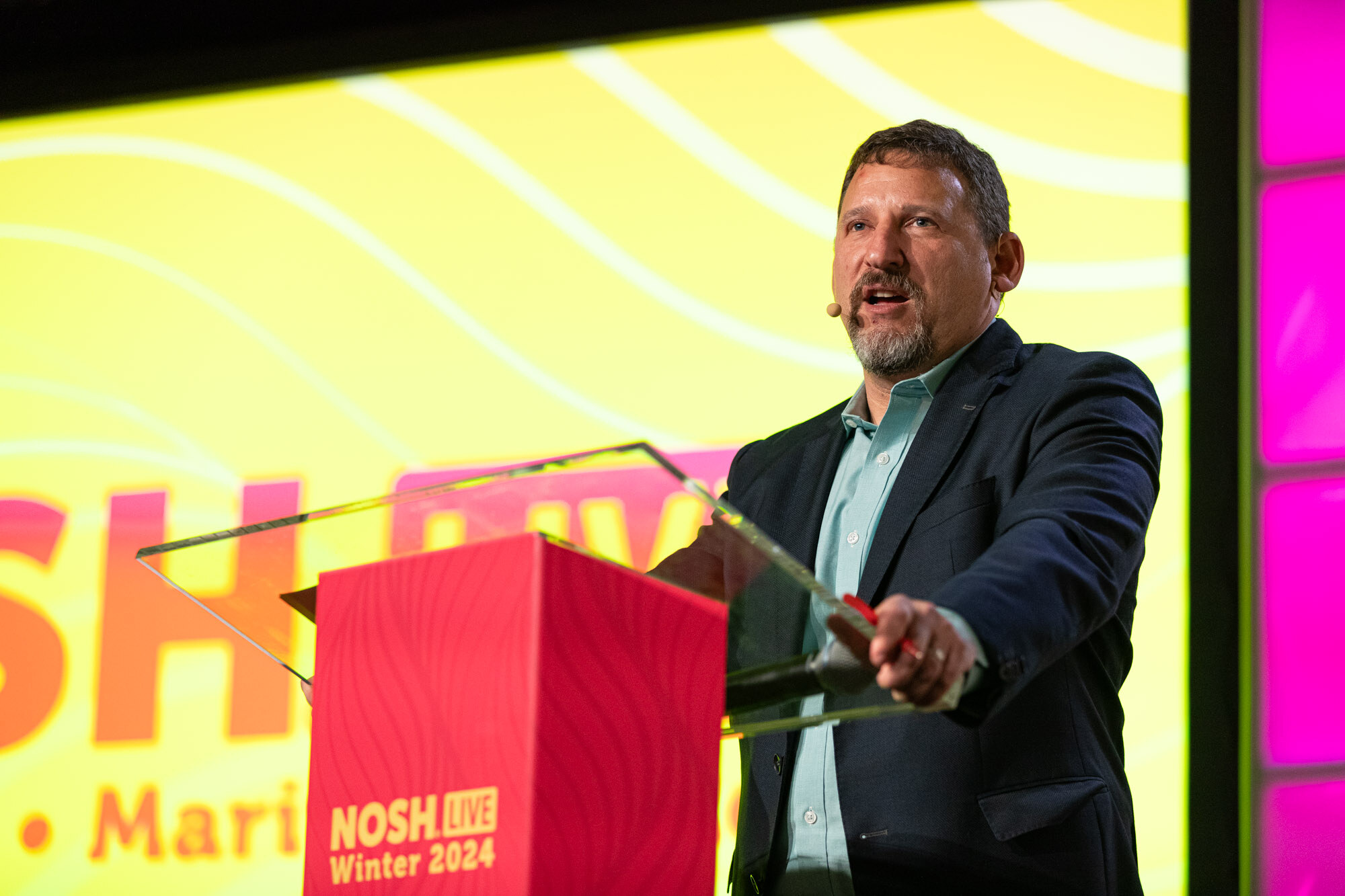 A man with a gray beard in a navy blazer and teal shirt holding a red pen at a red podium.