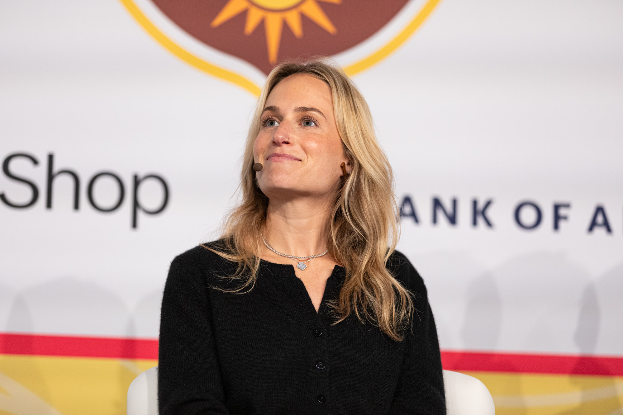 A woman with long blond hair in a black cardigan seated on stage in front of a sponsor wall.