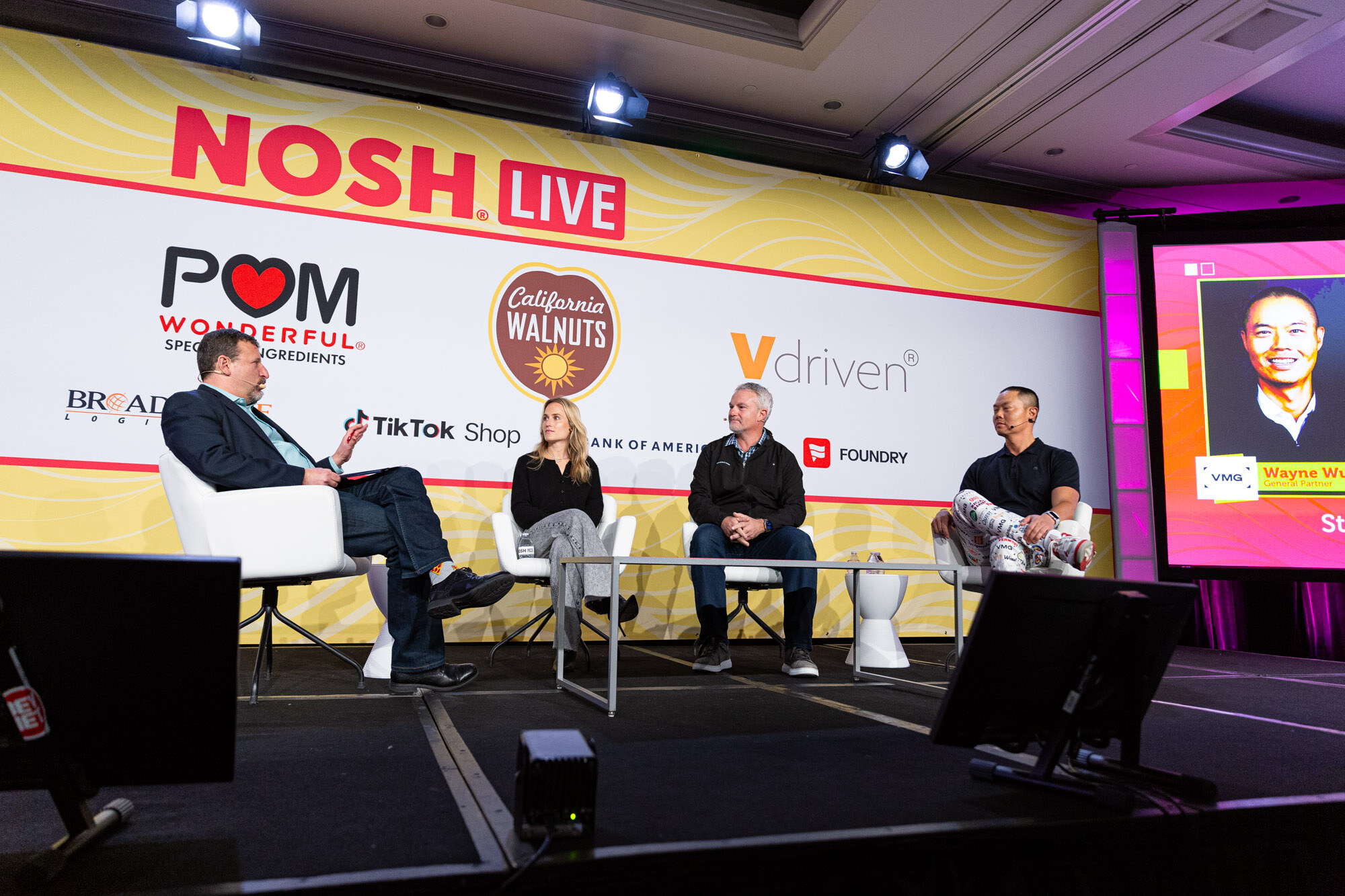 Wide view of four people seated in white chairs on a stage with a yellow Nosh Live backdrop and a screen showing a man's portrait.