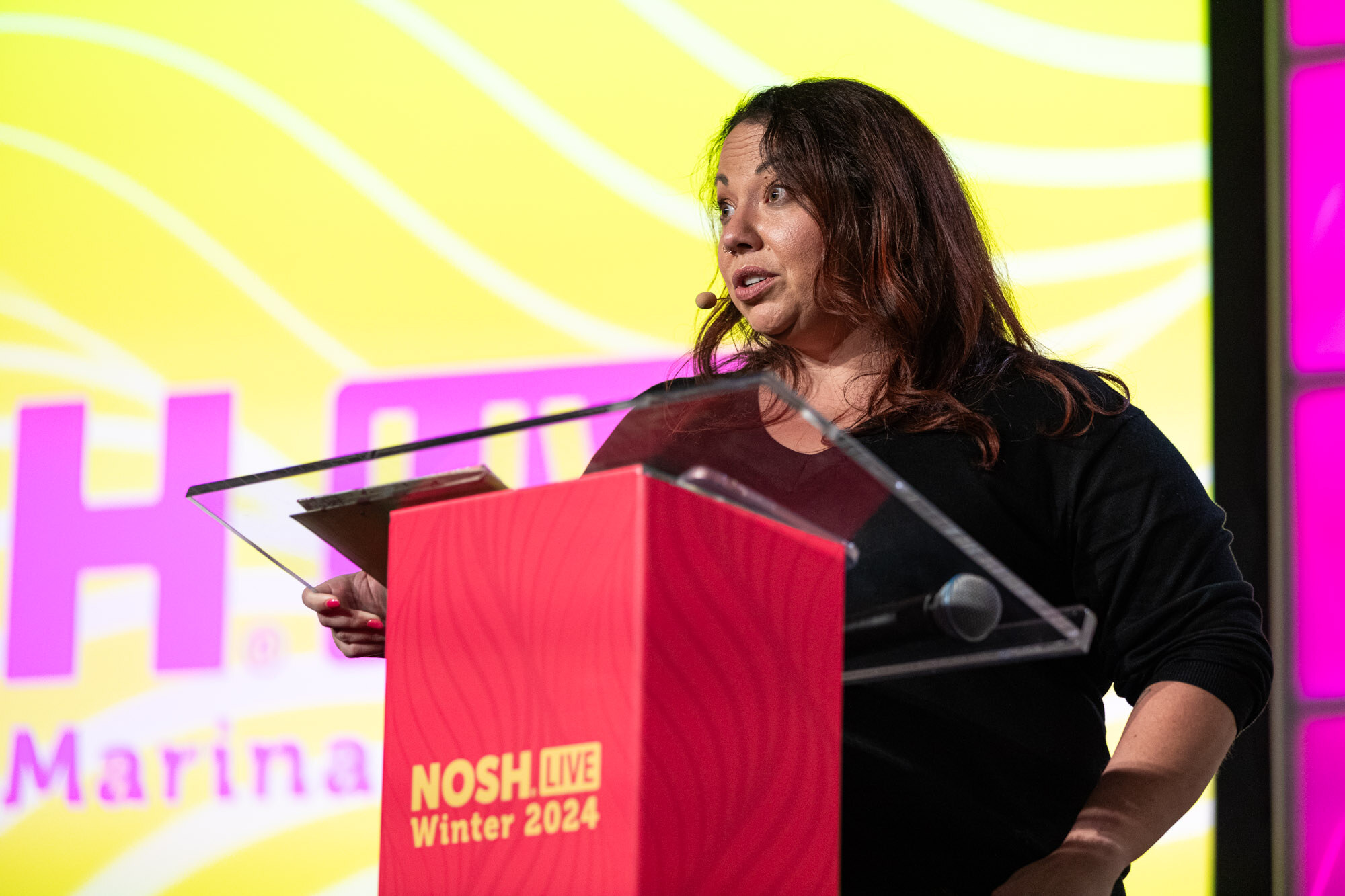 A woman with dark wavy hair in a black top at a red podium with a yellow Nosh Live screen behind her.