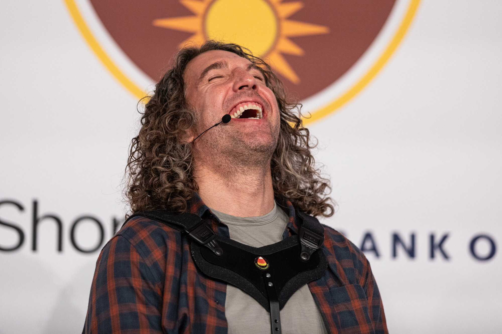 A man with long curly hair laughing with his head back, wearing a plaid shirt over a gray back-support brace.