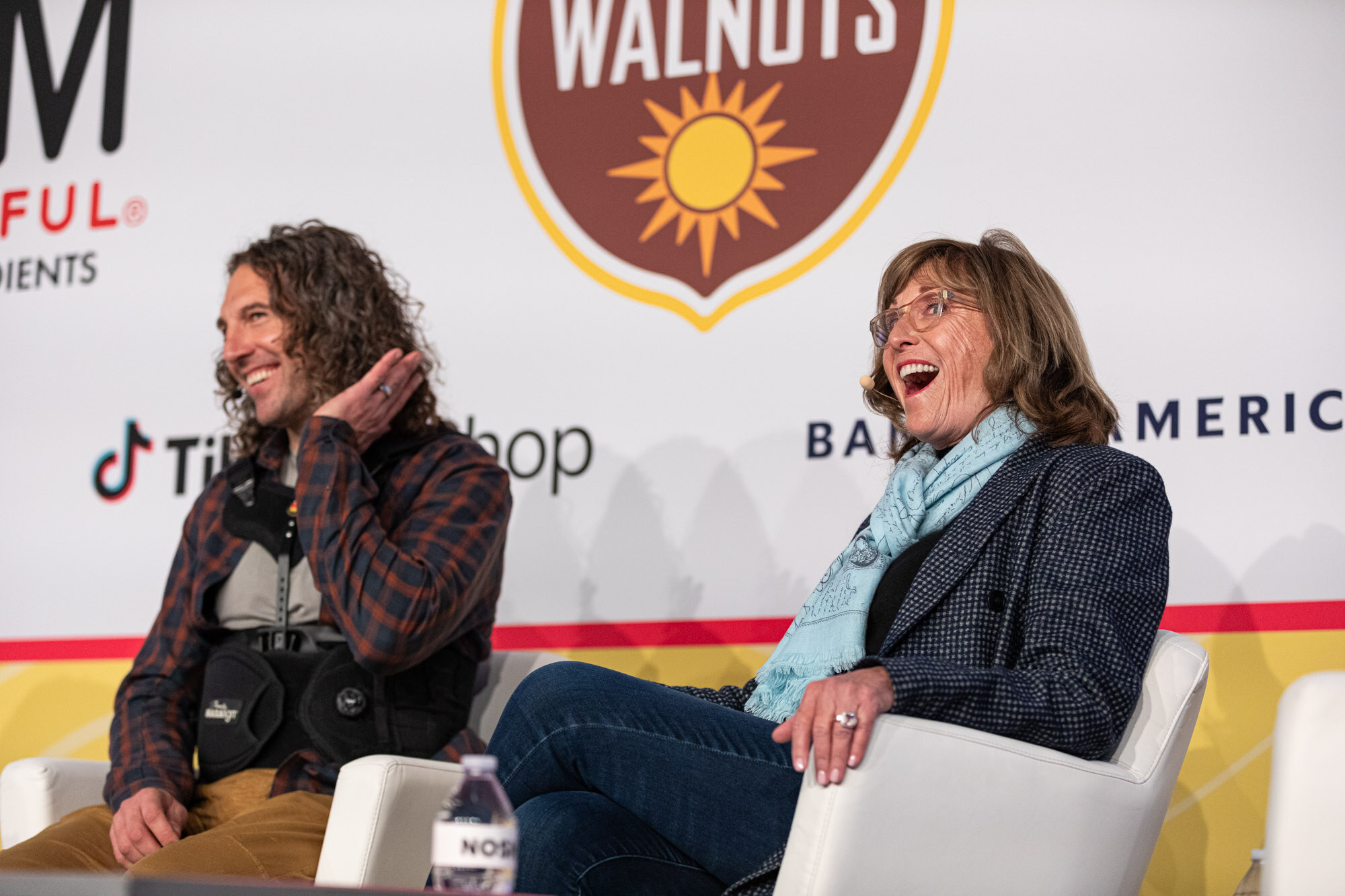 A woman in a light-blue scarf looking amazed beside a long-haired man in a plaid shirt, both seated on stage.