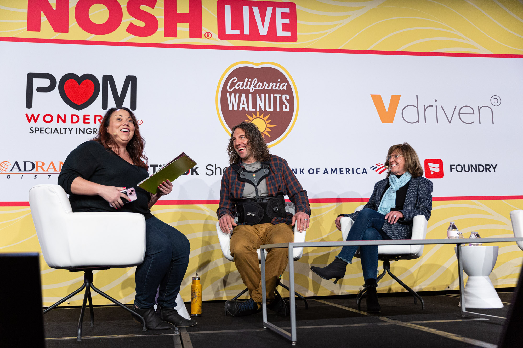 Three people seated on a stage in front of a Nosh Live backdrop, laughing, one holding a green folder.