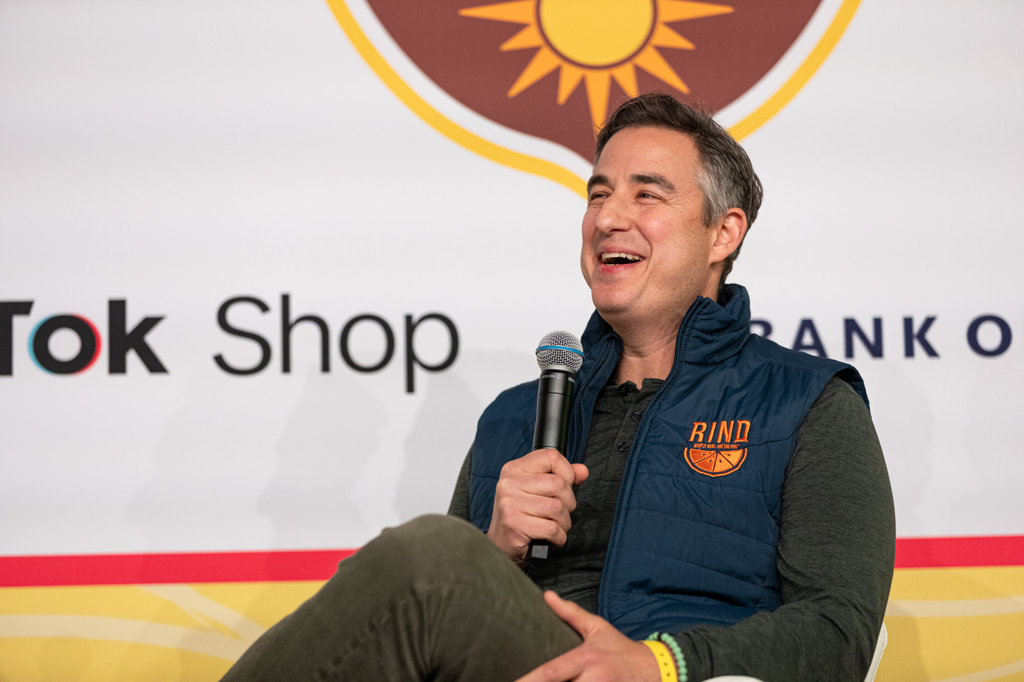 A smiling man in a navy puffer vest marked Rind, holding a microphone and seated on stage.