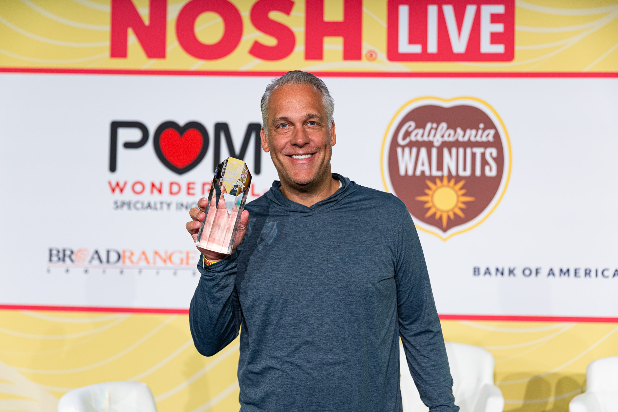 A smiling gray-haired man in a blue hoodie holding a glass trophy in front of a sponsor backdrop.