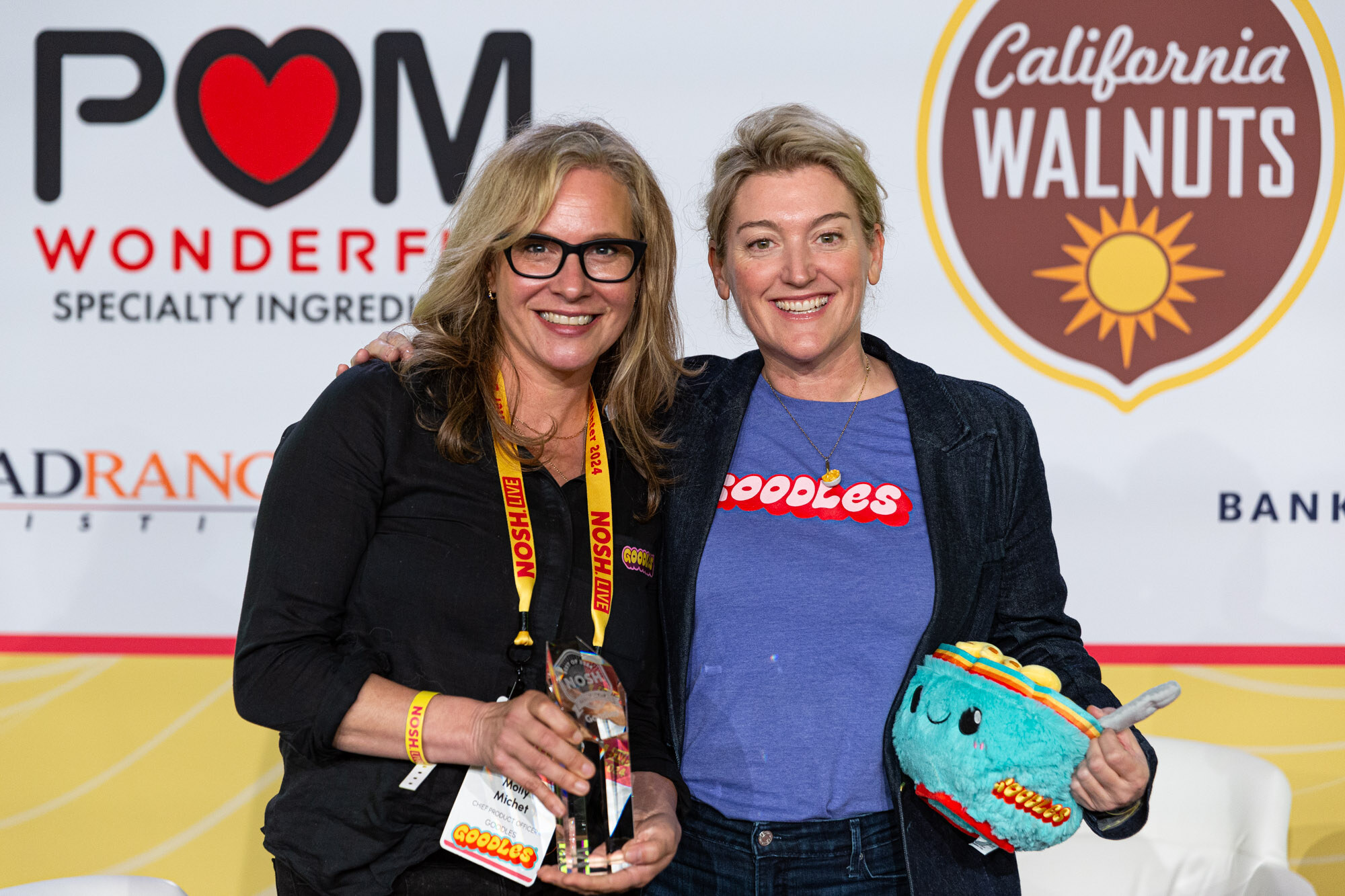 Two women, one wearing a blue Goodles t-shirt and holding a turquoise plush bowl, standing together in front of a sponsor wall.