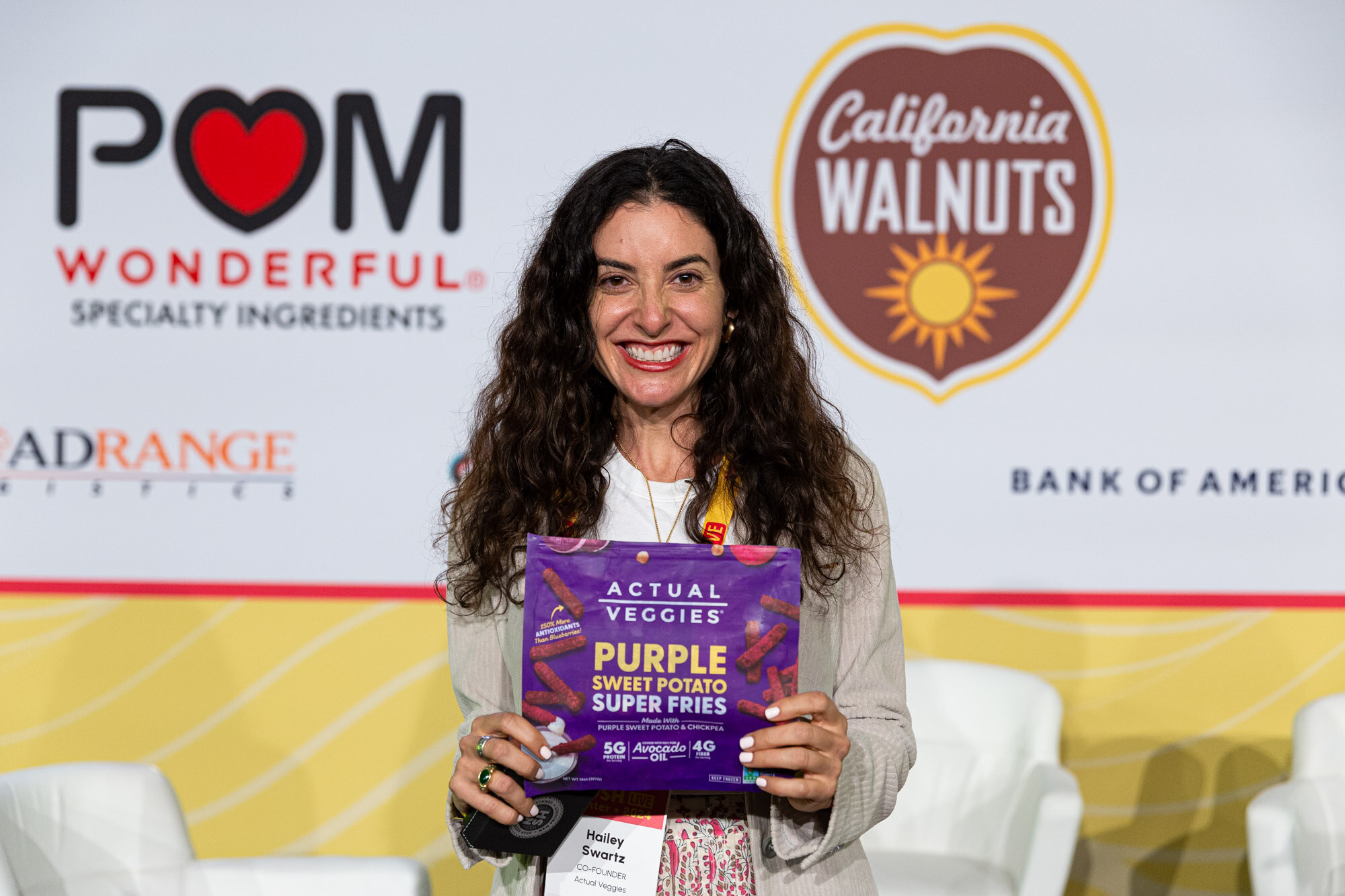A woman with curly dark hair holding a purple bag labeled Actual Veggies Purple Sweet Potato Super Fries.