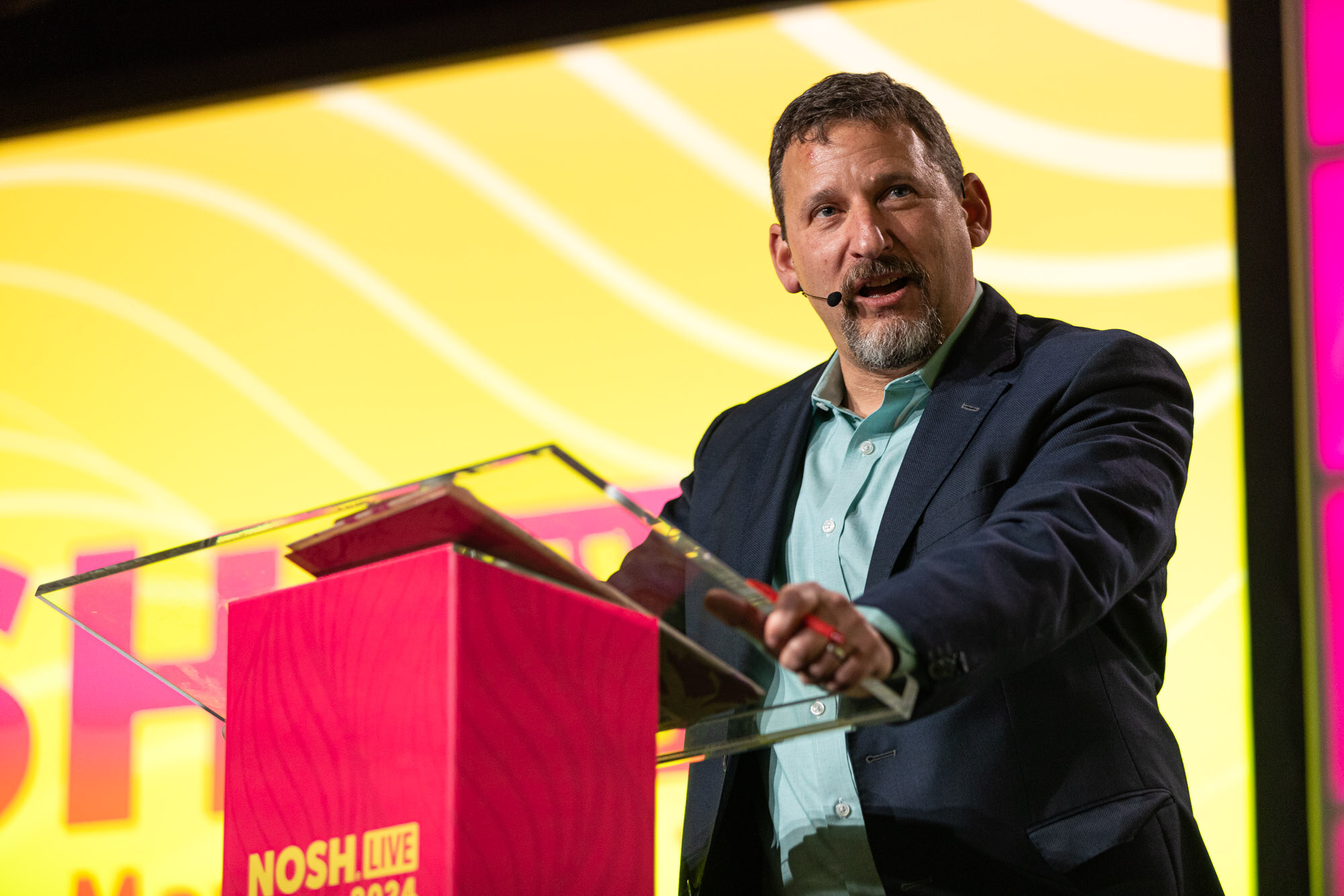 A man with a graying beard in a navy blazer at a red podium in front of a yellow screen.