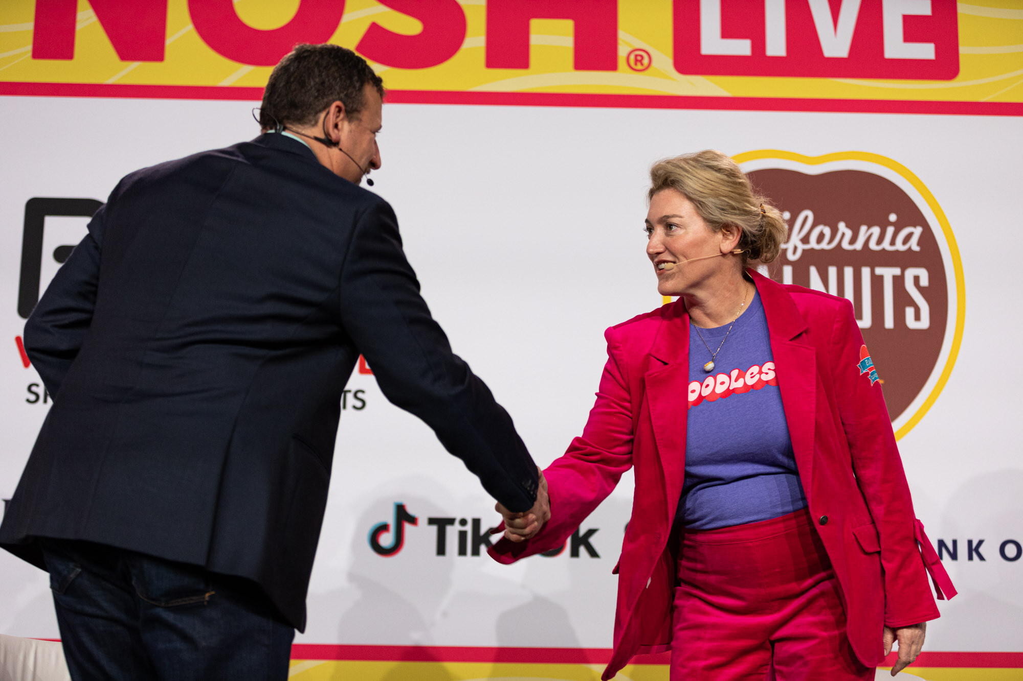 A man in a navy blazer shaking hands with a woman in a hot-pink suit and a blue Goodles t-shirt.
