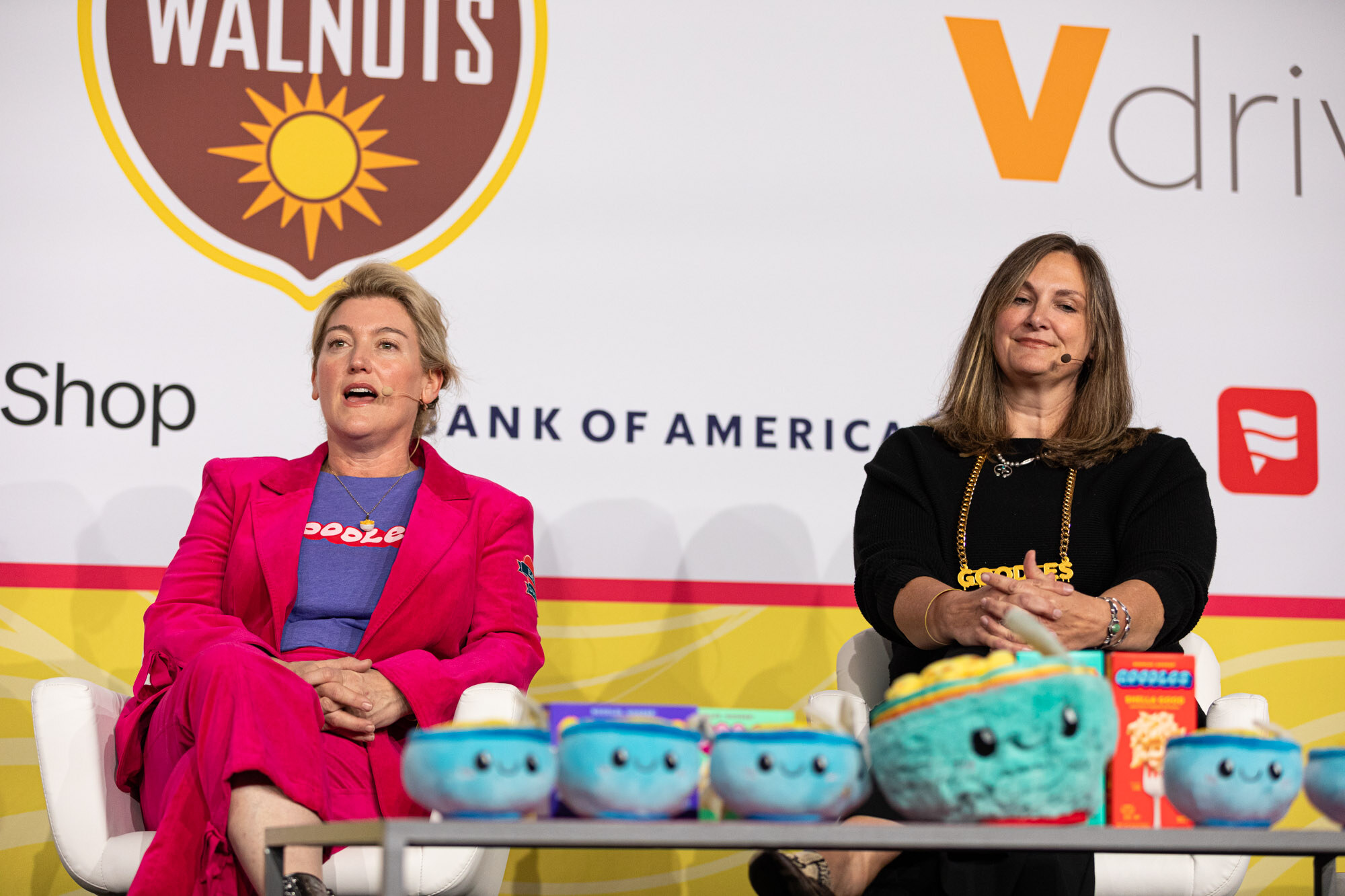 Two women seated on a stage behind a table lined with blue plush bowls and a turquoise plush mascot.