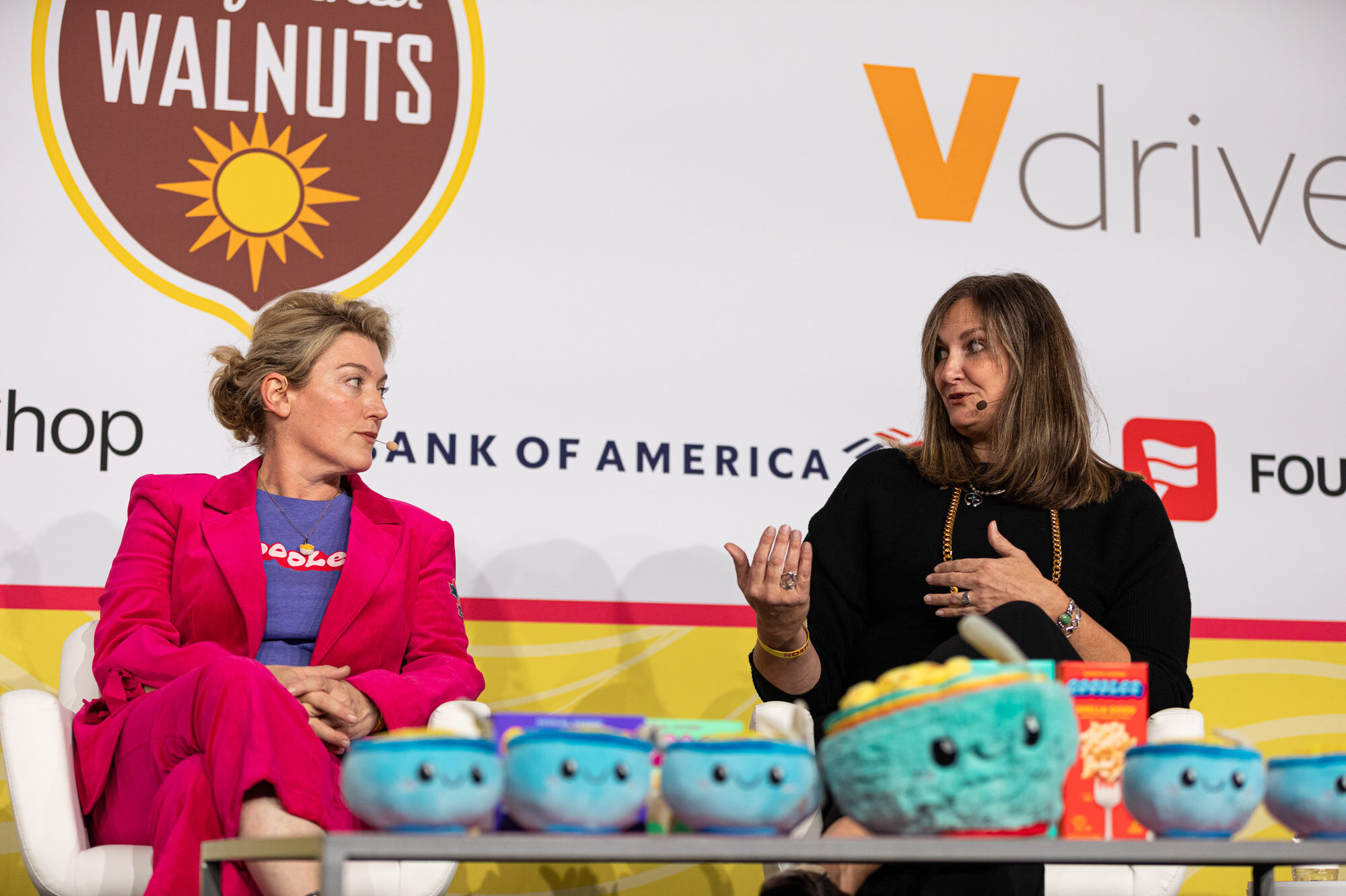 A woman in a pink suit and another in black with a gold necklace sit behind blue plush bowls and talk.