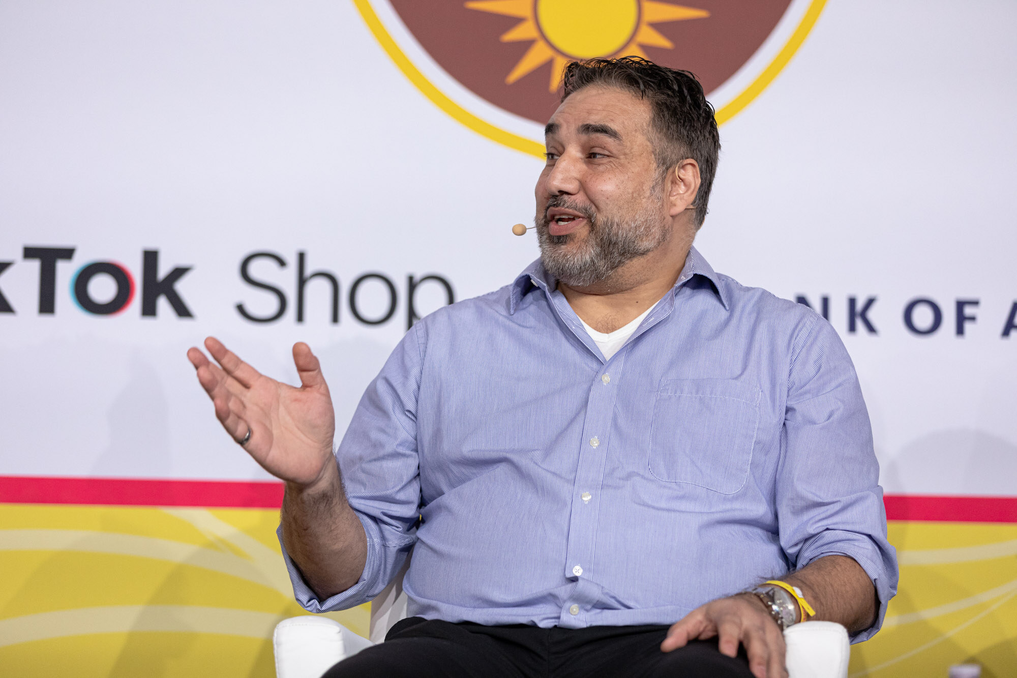 A bearded man in a periwinkle button-down shirt gesturing with one hand while seated on stage.