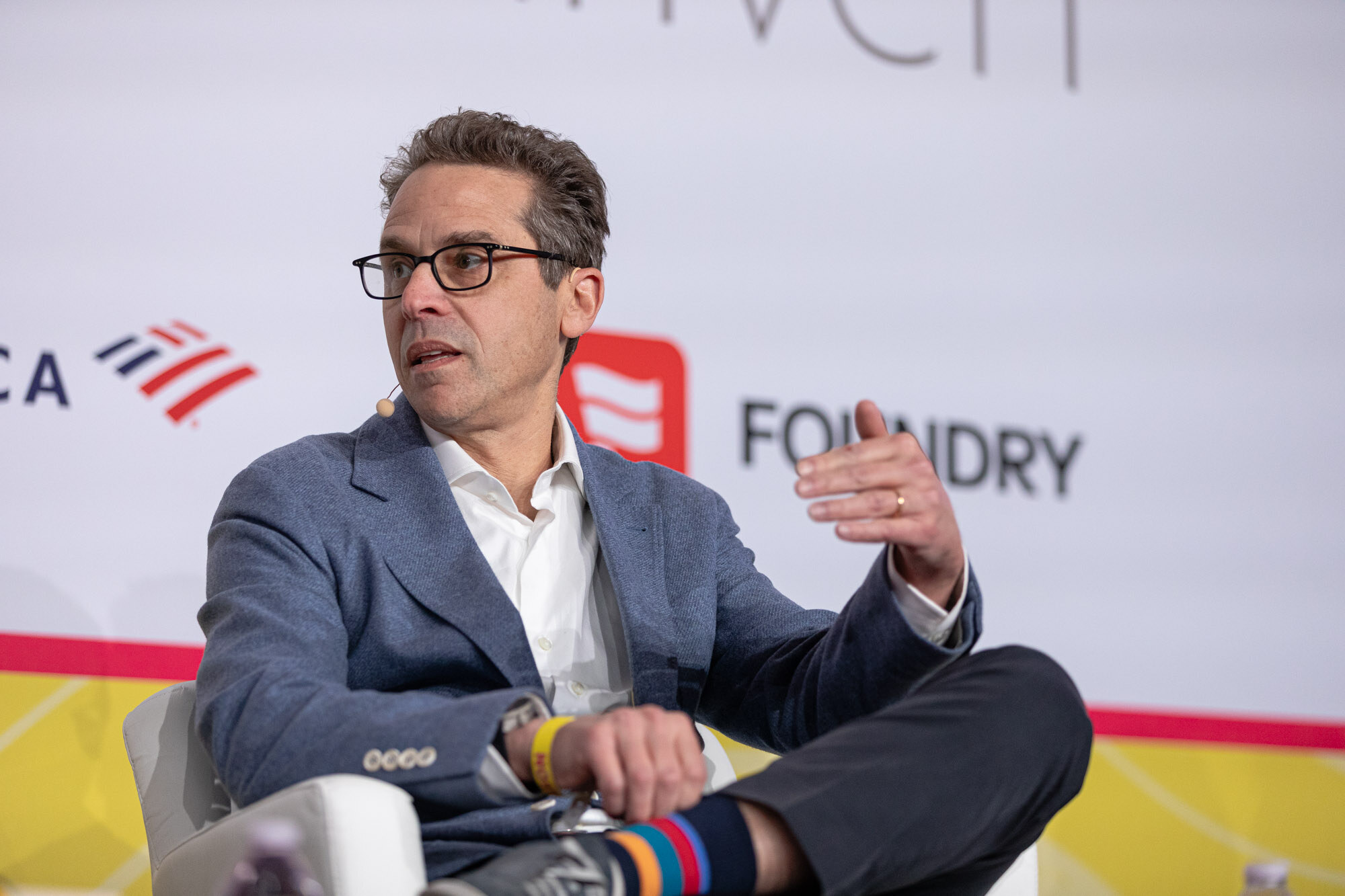 A man in dark glasses, a blue blazer and rainbow-striped socks, seated with crossed legs and gesturing.